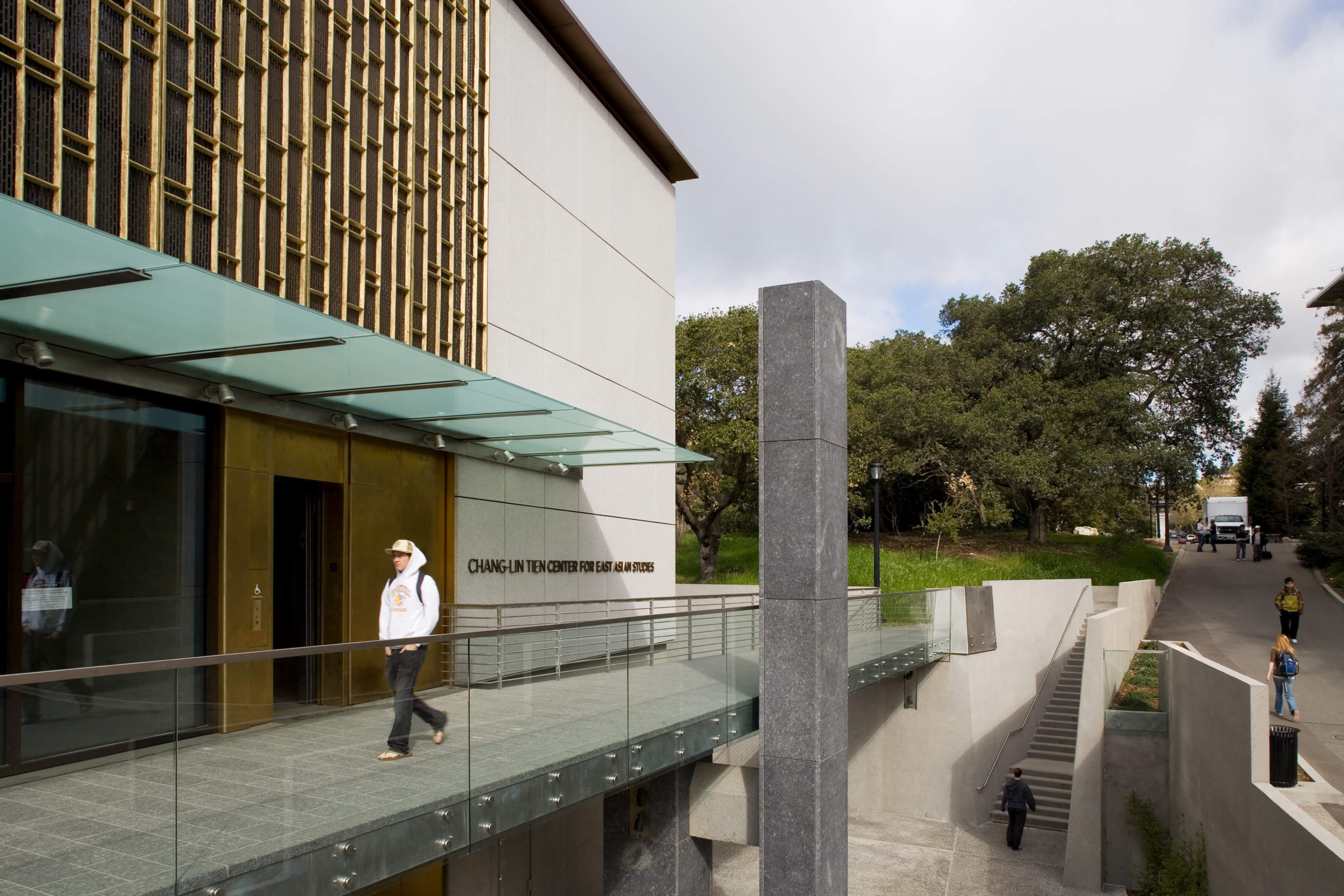 Exterior view of the Chang-Lin Tien Center for East Asian Studies, showing patterned bronze screens, a glass entry canopy, and a raised terrace connected to stairs and walkways along the sloped campus site.