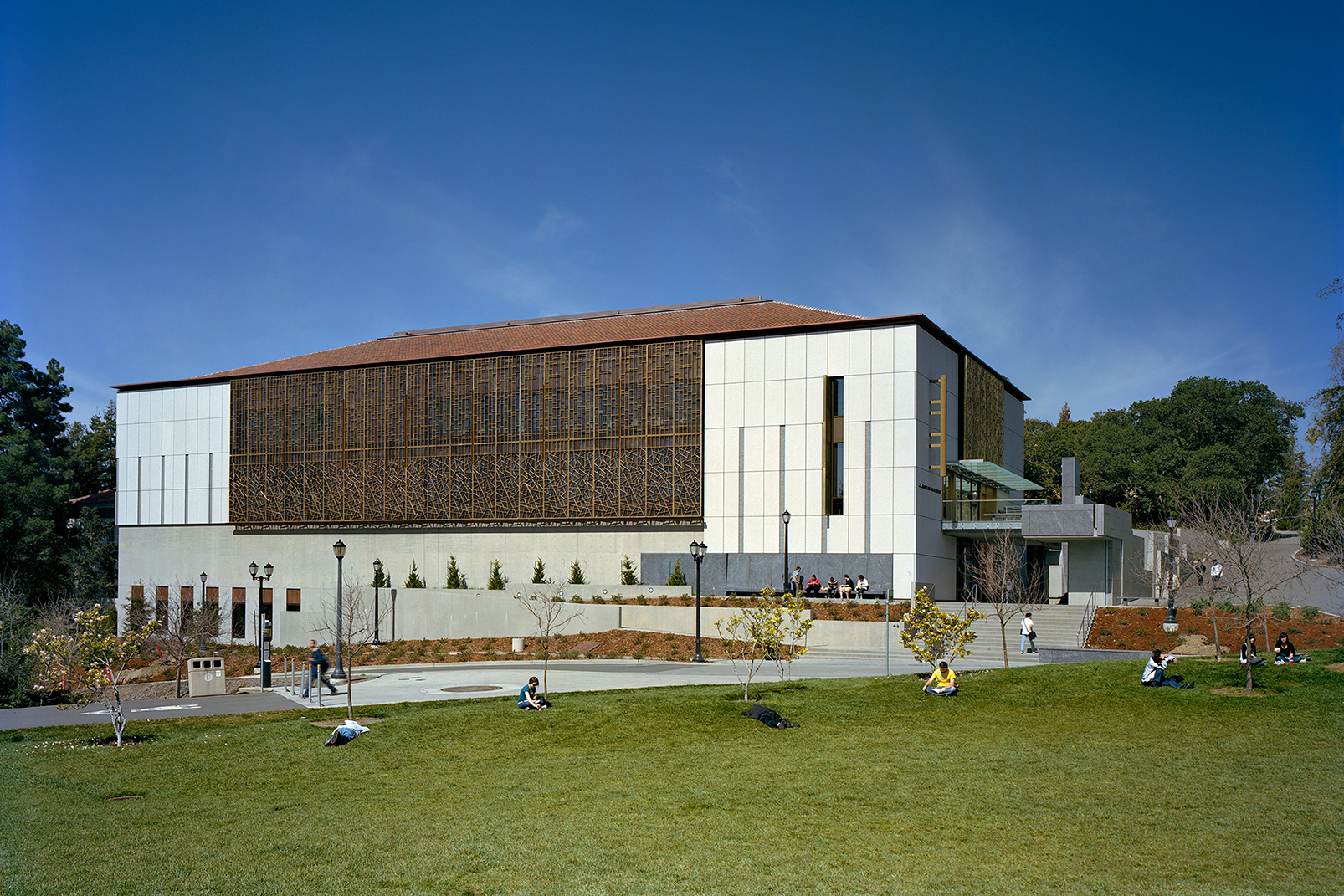 A granite-clad campus library with patterned bronze screens sits at the edge of a large lawn under a clear blue sky.