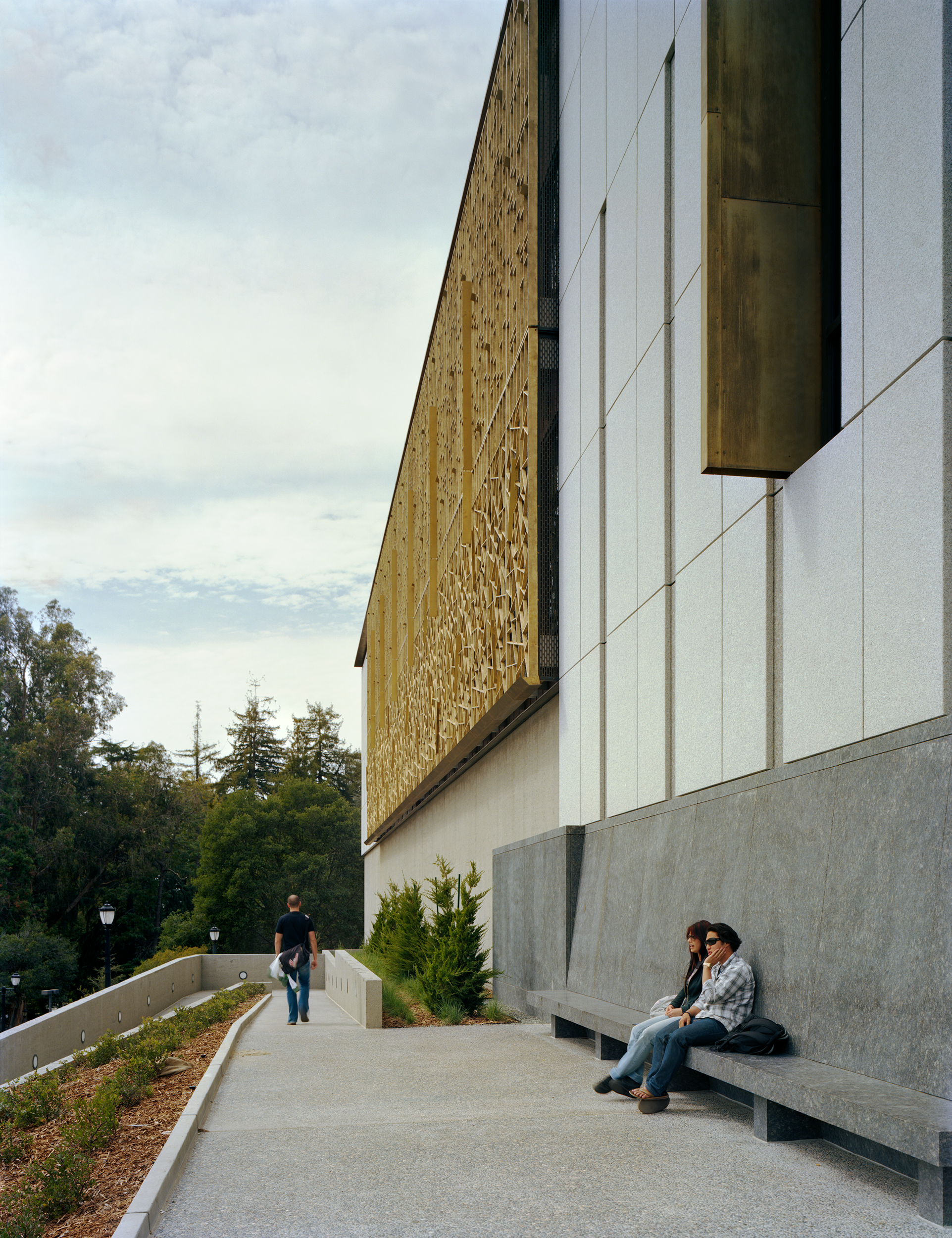 Exterior walkway along the East Asian studies library, with granite cladding, patterned bronze screens, landscaped edges, and built-in stone seating along the sloped campus site.