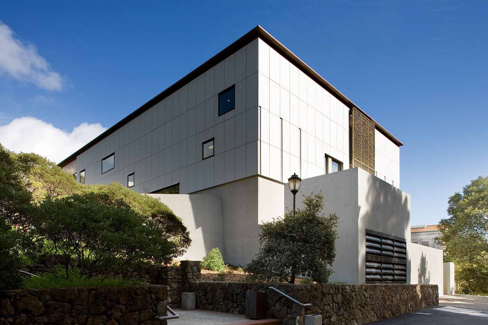 Exterior view of a granite-clad East Asian studies library set into a sloped campus site, with patterned bronze screens, small punched windows, concrete retaining walls, stone landscaping, and a clear blue sky overhead.
