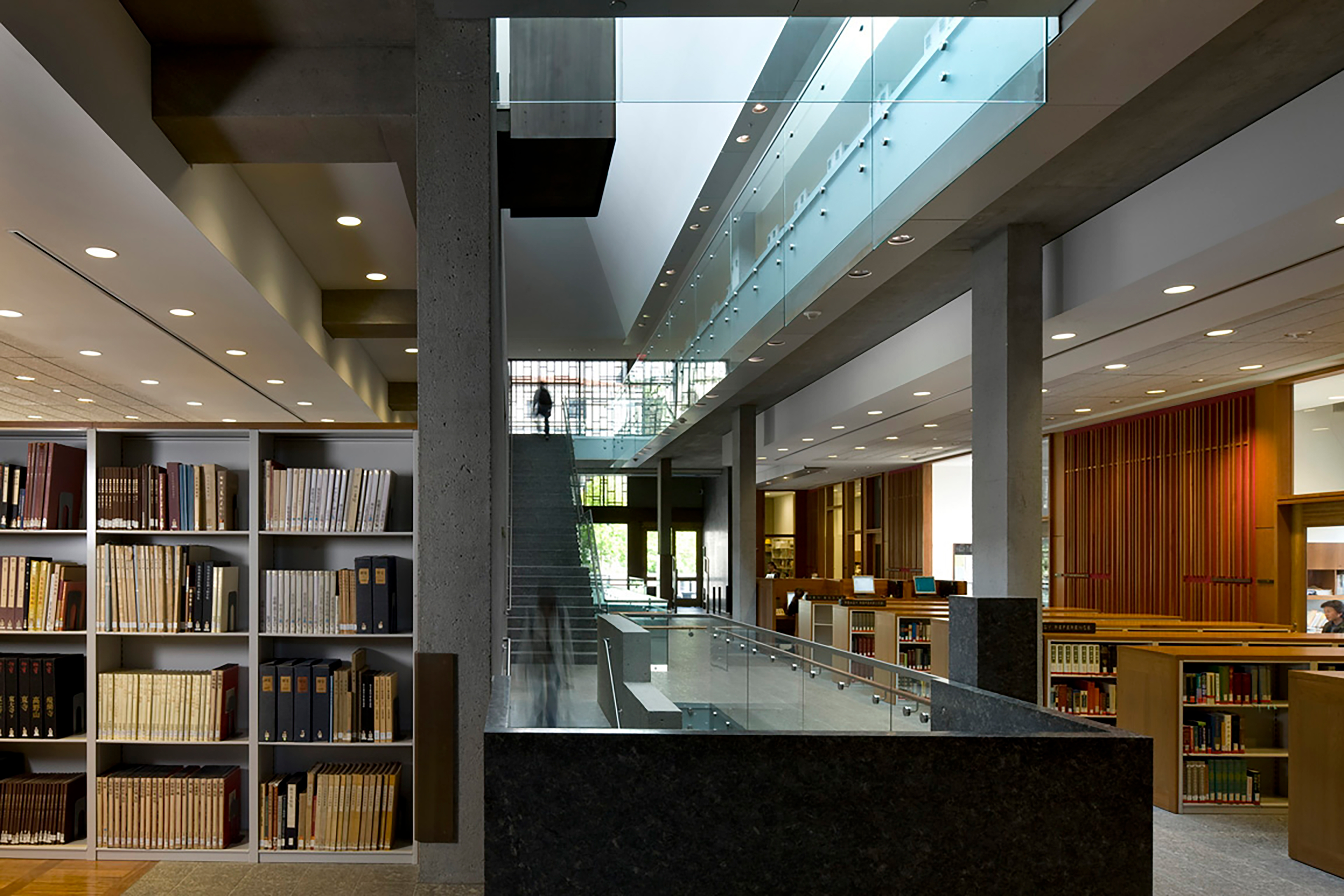 Interior view of a multi-level library with book stacks, glass railings, concrete columns, and a central stair that draws filtered daylight through the building.