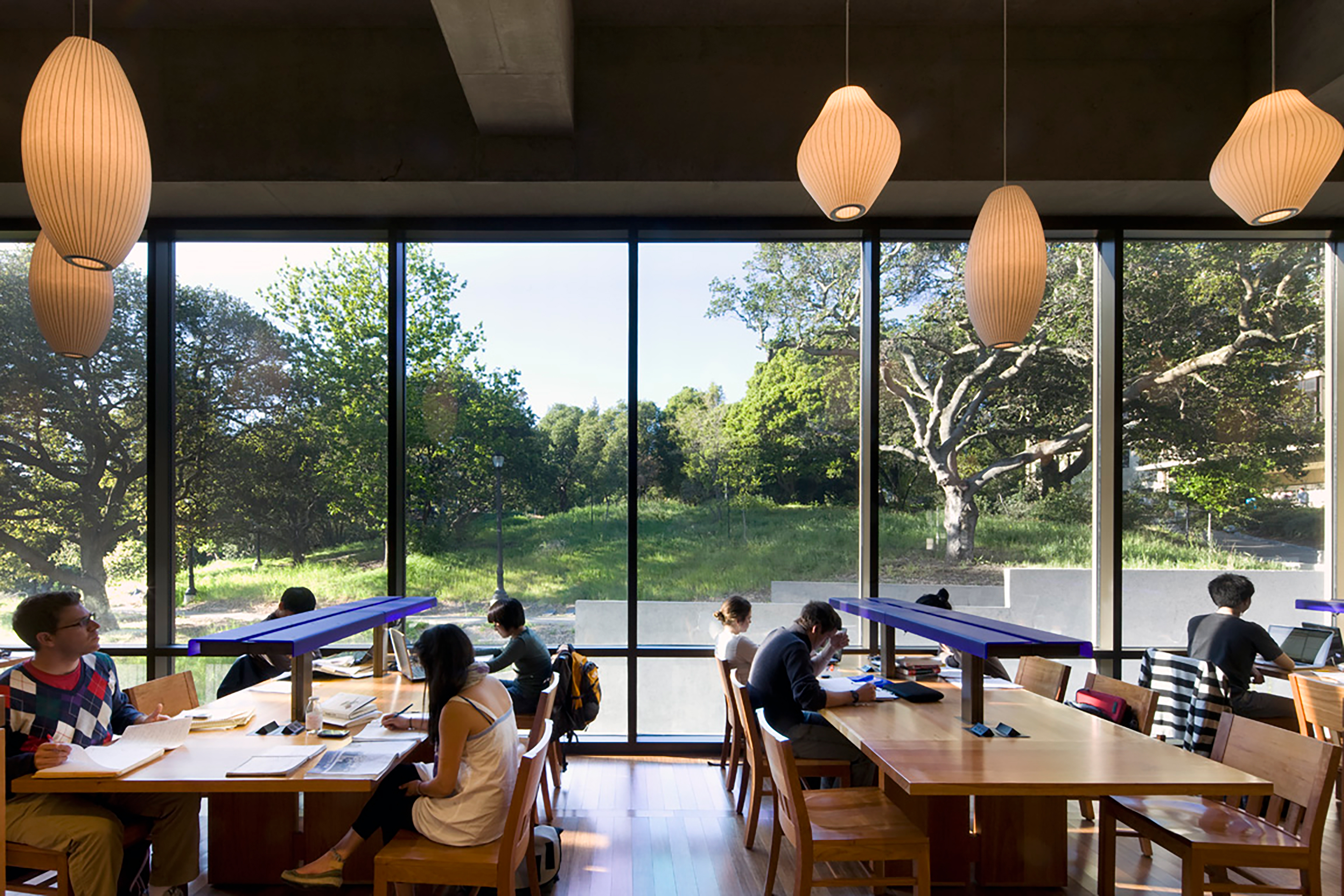 Students study at wood reading tables beside full-height windows overlooking a landscaped campus, with warm pendant lights suspended above.