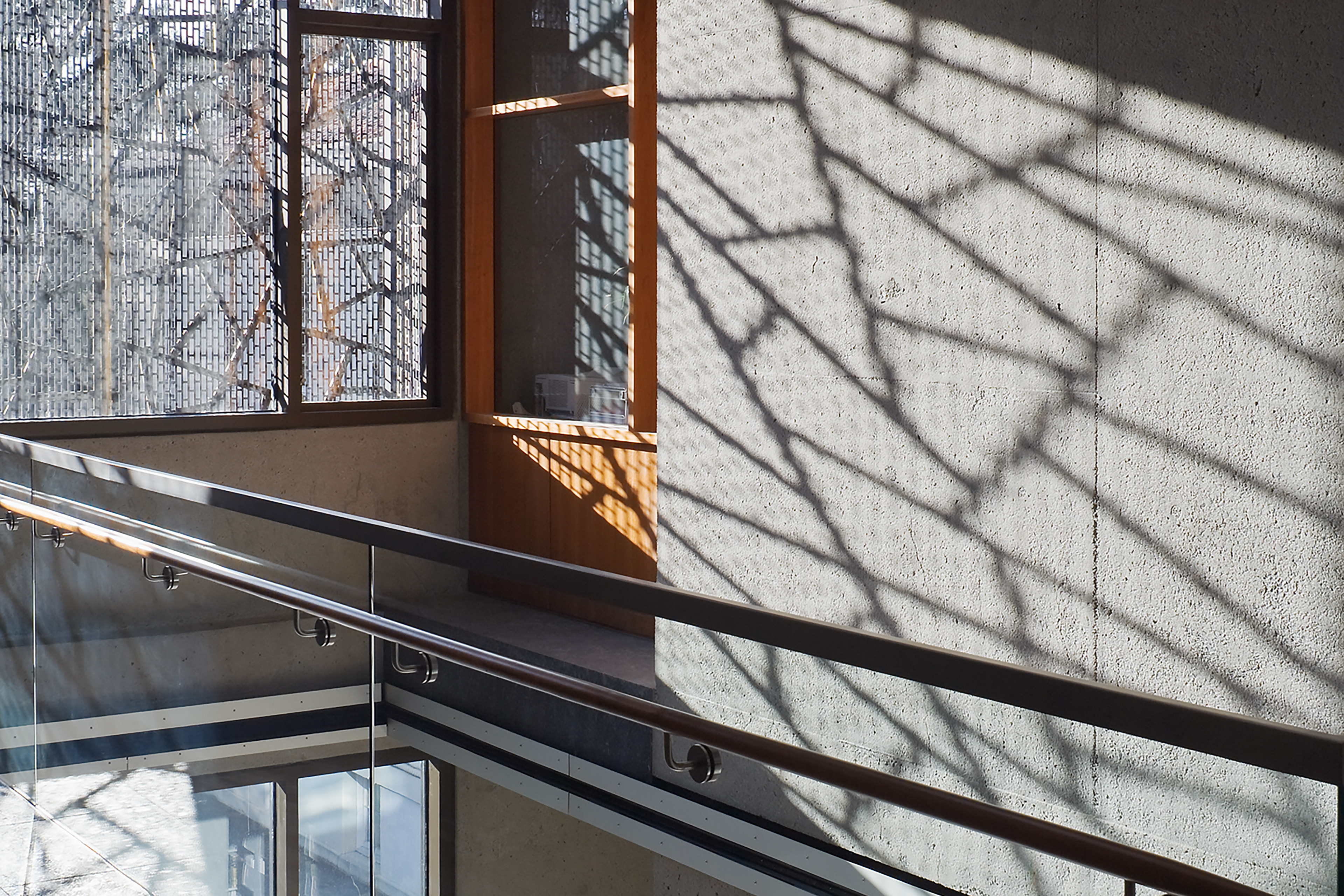 Interior walkway with glass railings and concrete walls, where patterned exterior screens filter sunlight and cast intricate shadows across the space.