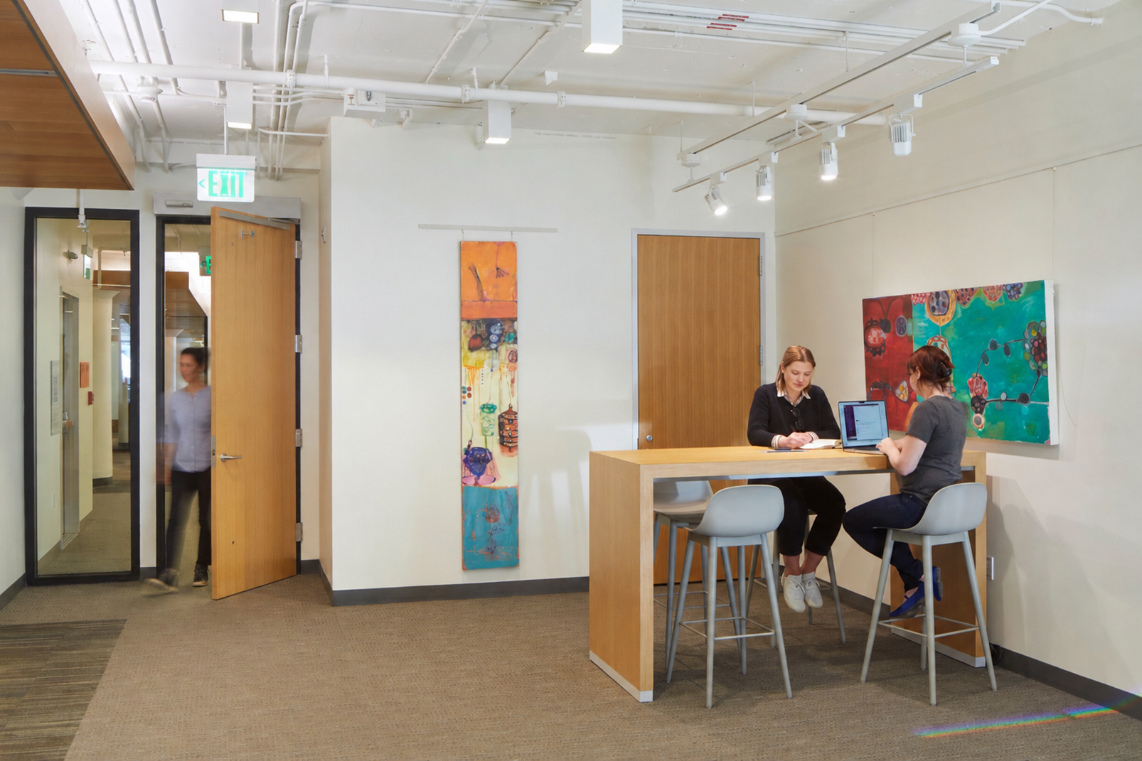 Modern office breakout area with a high wood table and gray stools beneath exposed white ceiling systems. Two women work together at the table, one writing in a notebook and the other using a laptop, while a motion-blurred person walks through an open doorway beneath an illuminated exit sign. Colorful abstract artwork adds contrast to the otherwise neutral interior.