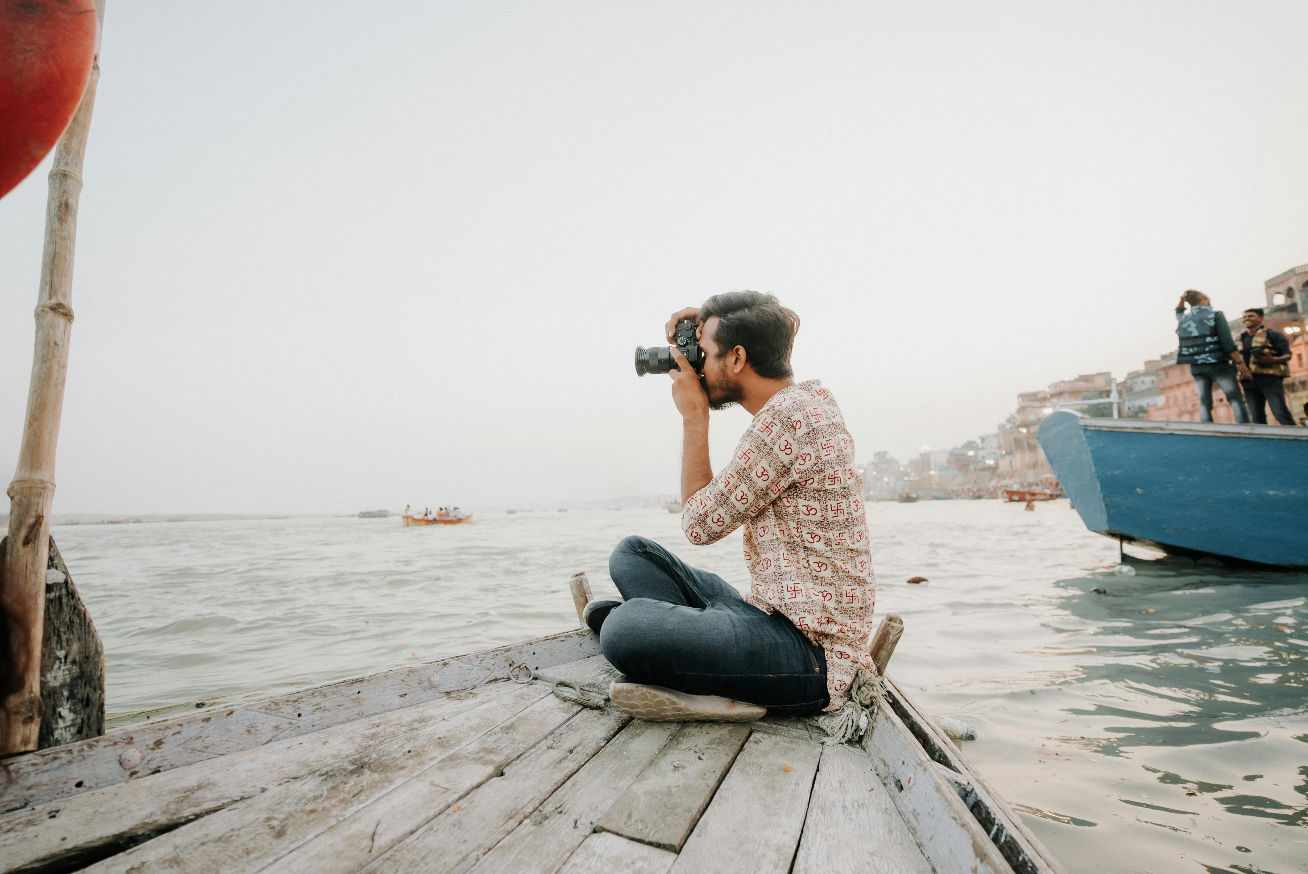 Man sitting on a dock taking a photo with a digital camera.