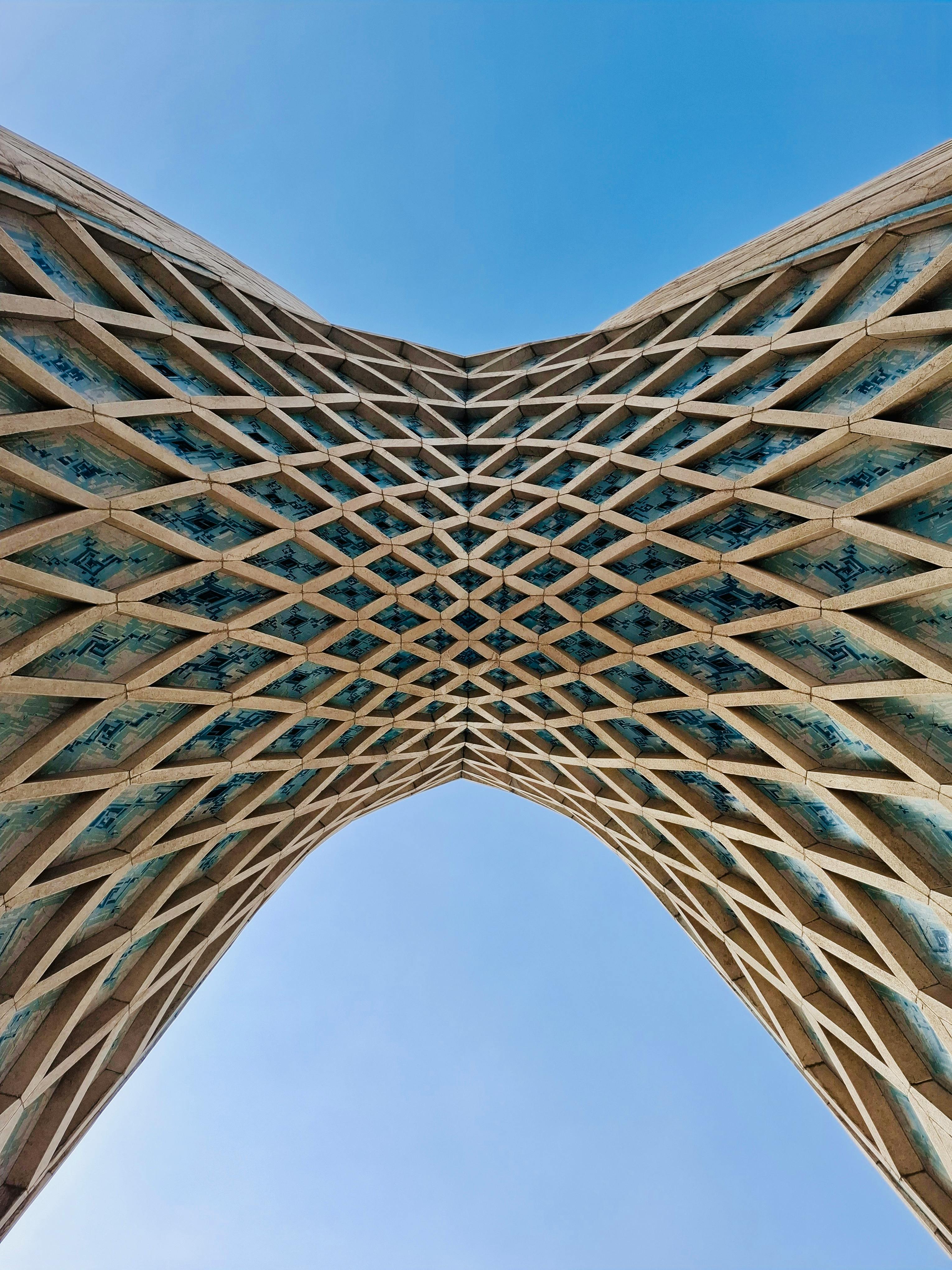 The underside of a criss-cross patterned architectural element, close enough to be abstract against a pale blue sky.