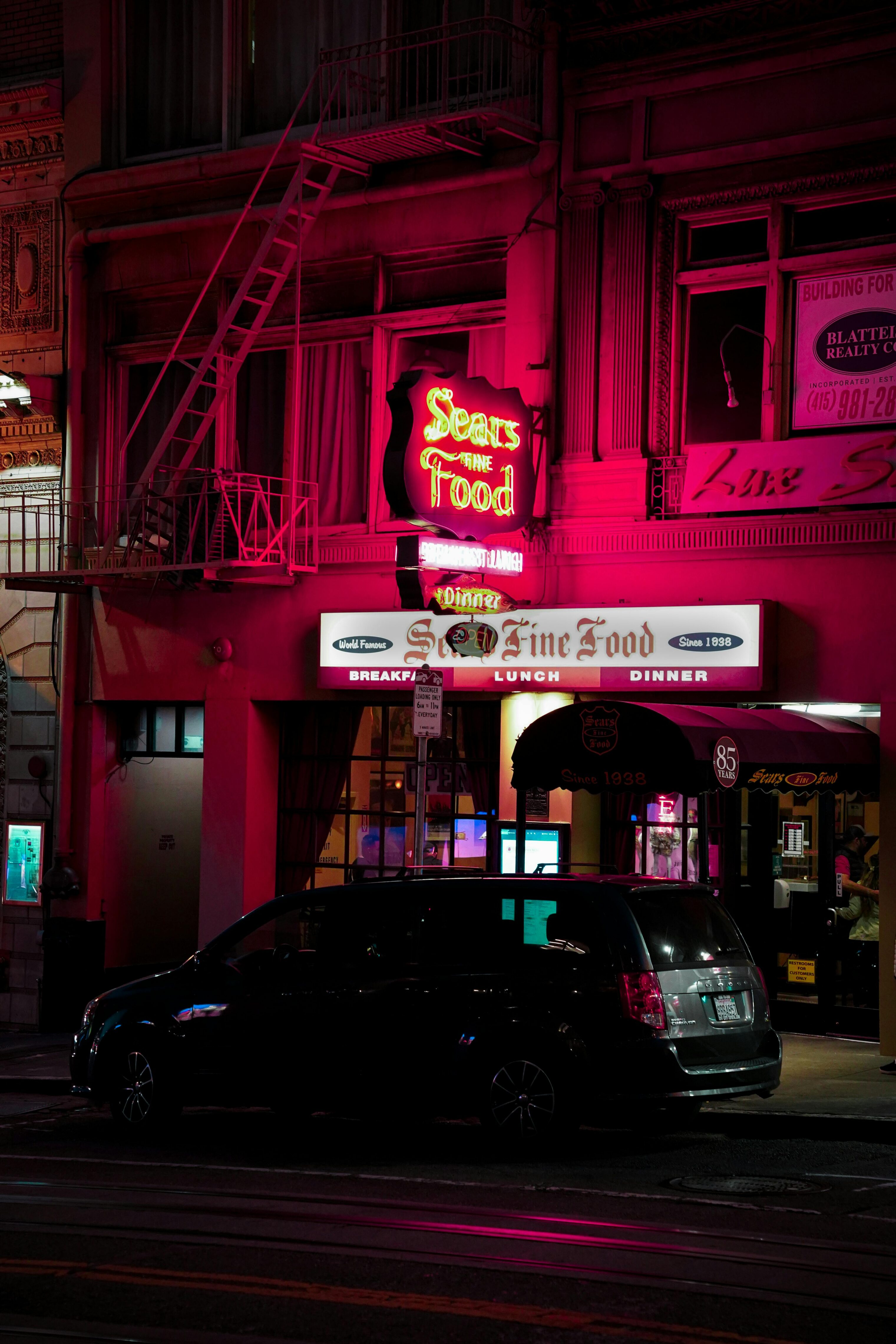 A nighttime city scene lit by a pink fluorescent sign advertising a sea food restaurant.