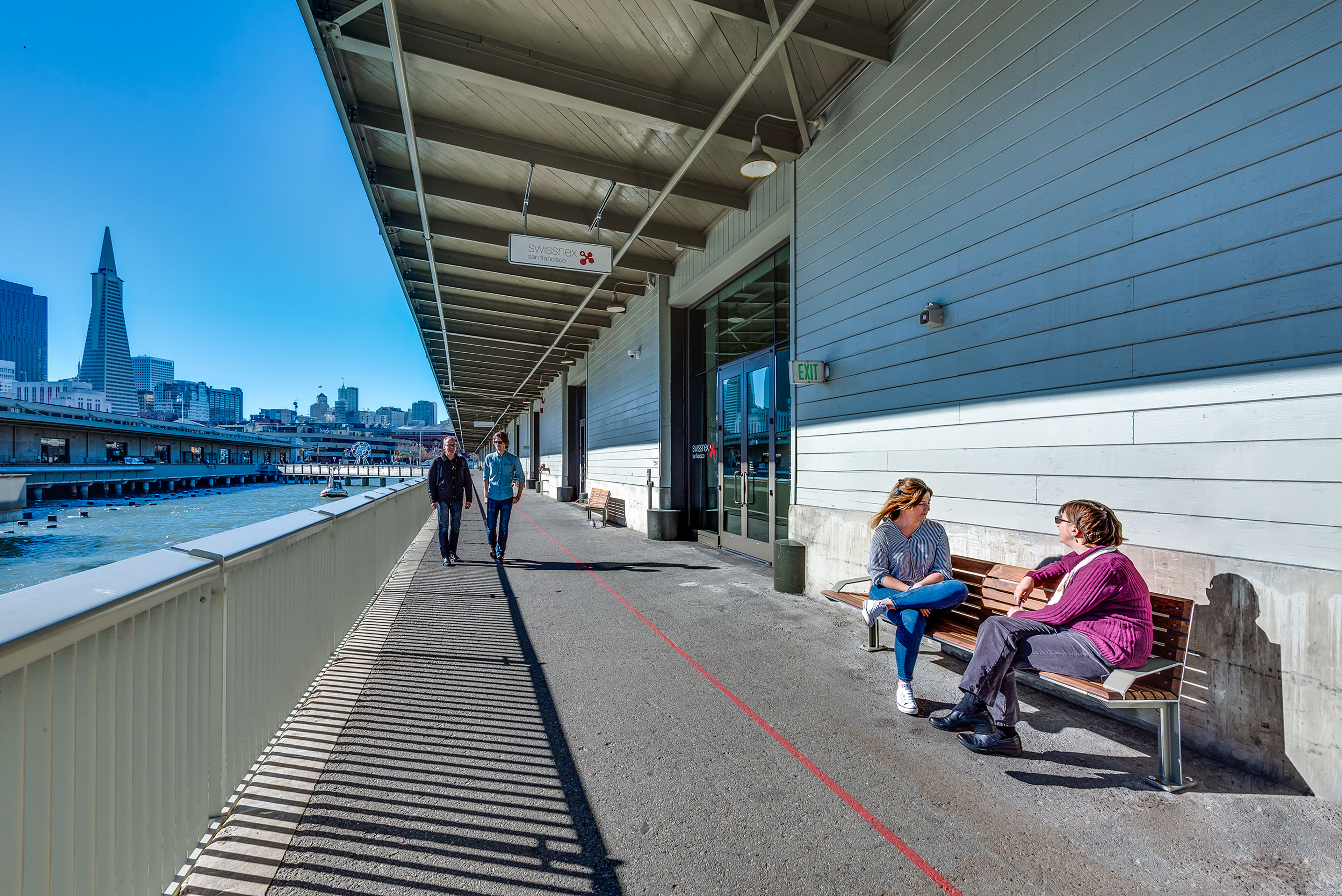 Sunny waterfront pier beside a gray-clad office building, with people walking along the railing and two people talking on a bench against the San Francisco skyline.