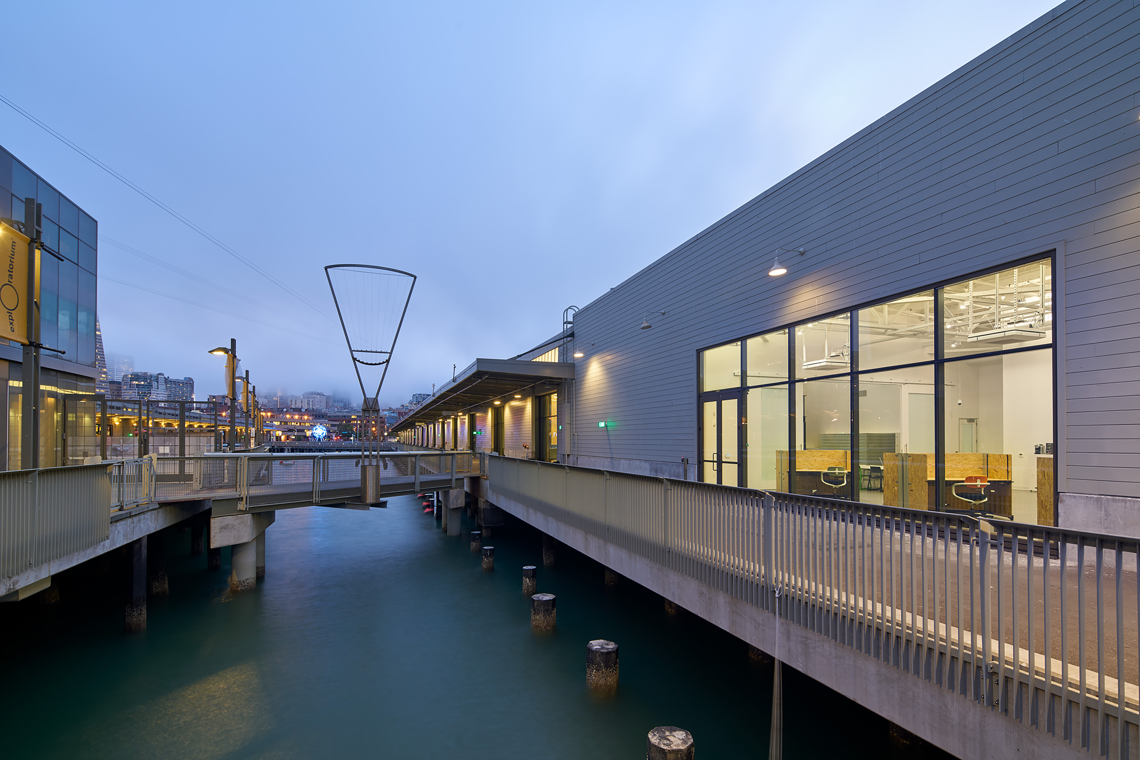 Dusk view of a waterfront office building and pier, with large windows revealing a bright workspace inside and city lights in the background.
