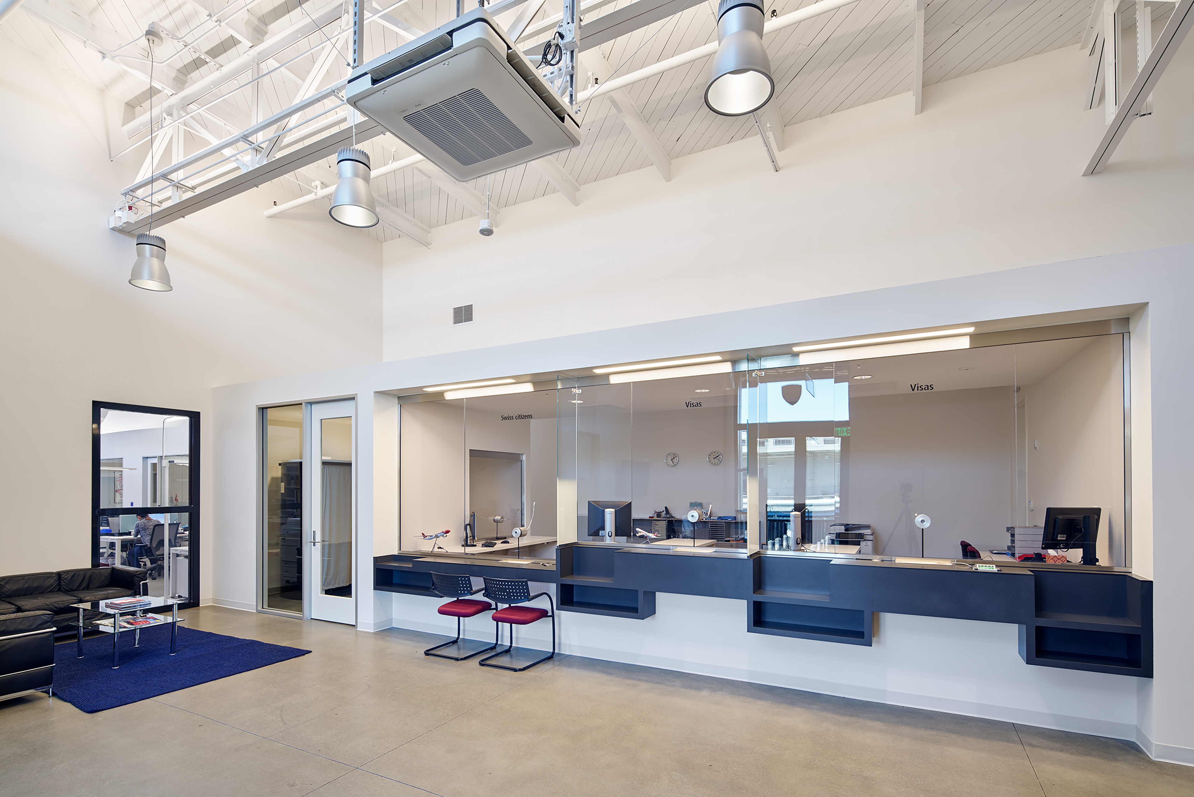 Modern consular service area with glass-fronted visa counters, a small waiting area, and an exposed white ceiling with industrial lighting.