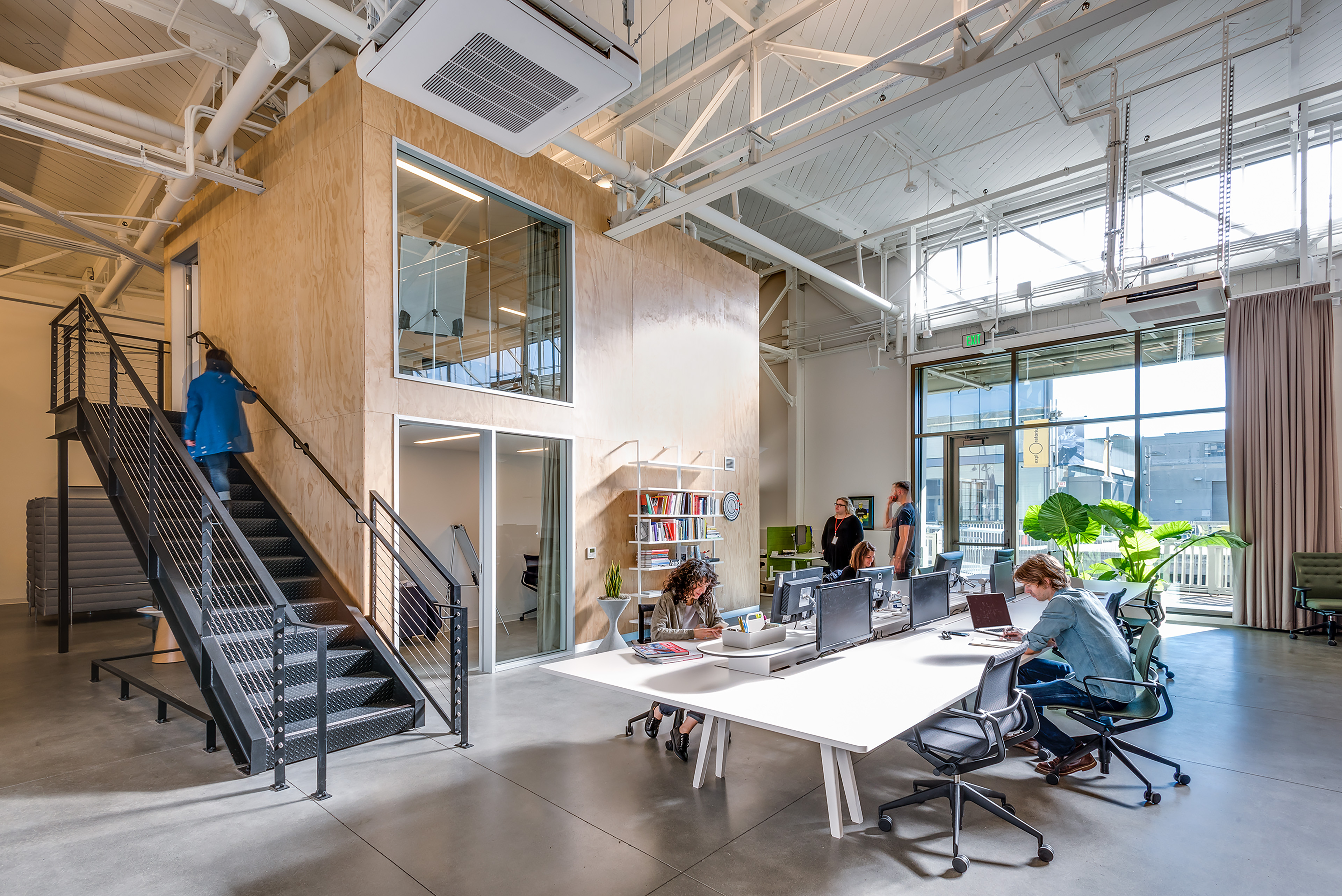 Open office with a plywood-clad mezzanine, exposed white trusses, black metal stair, shared desks, and people working beneath tall waterfront windows.