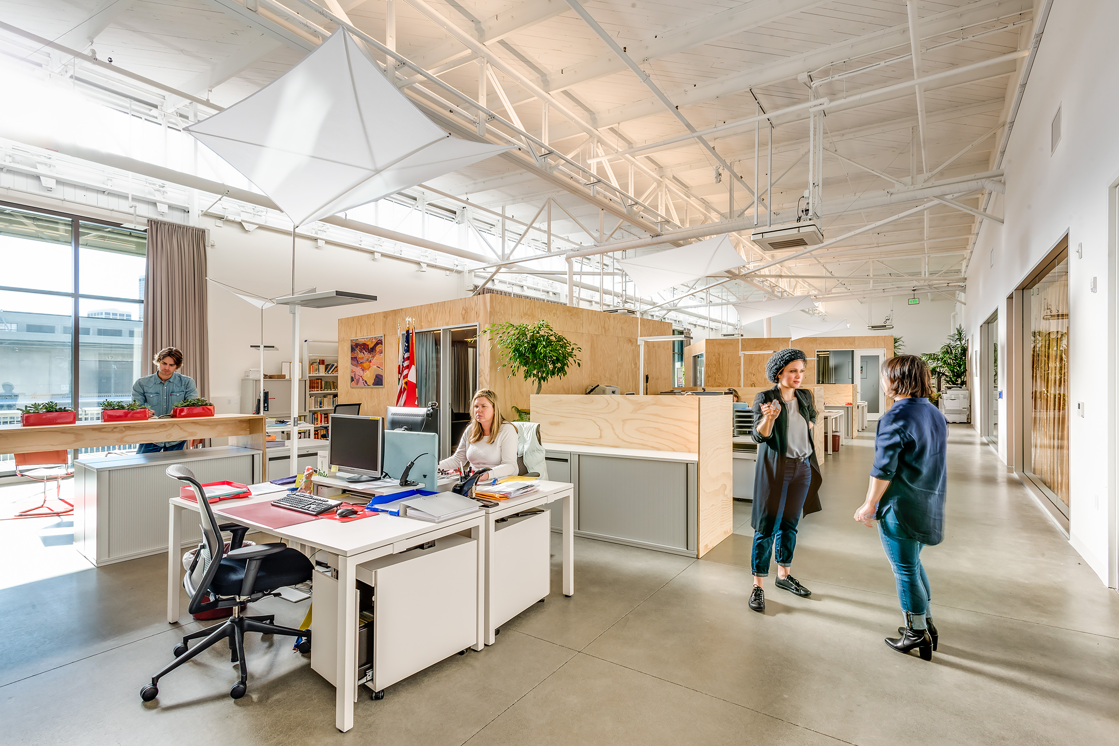 Bright open office with exposed white trusses, plywood partitions, desks, plants, and people working and having a conversation in the aisle.
