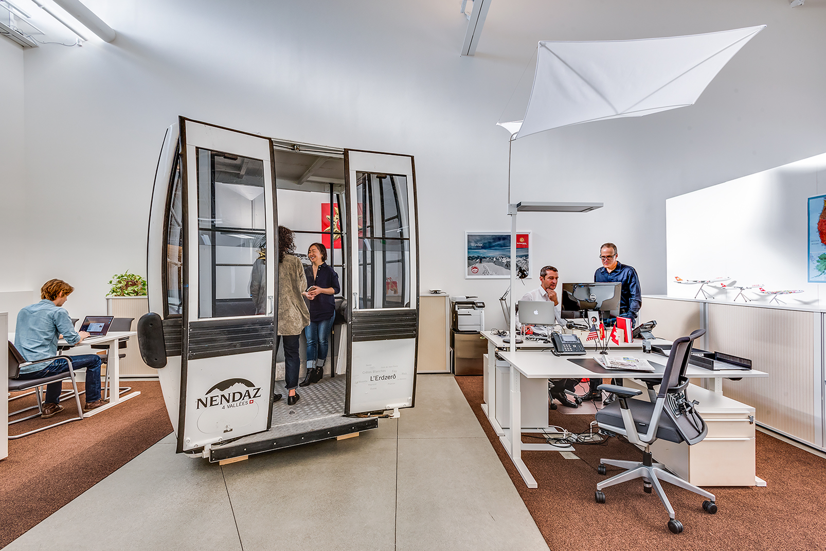 Open office with a repurposed gondola cabin used as a meeting room, surrounded by workstations, model airplanes, and people working and talking.