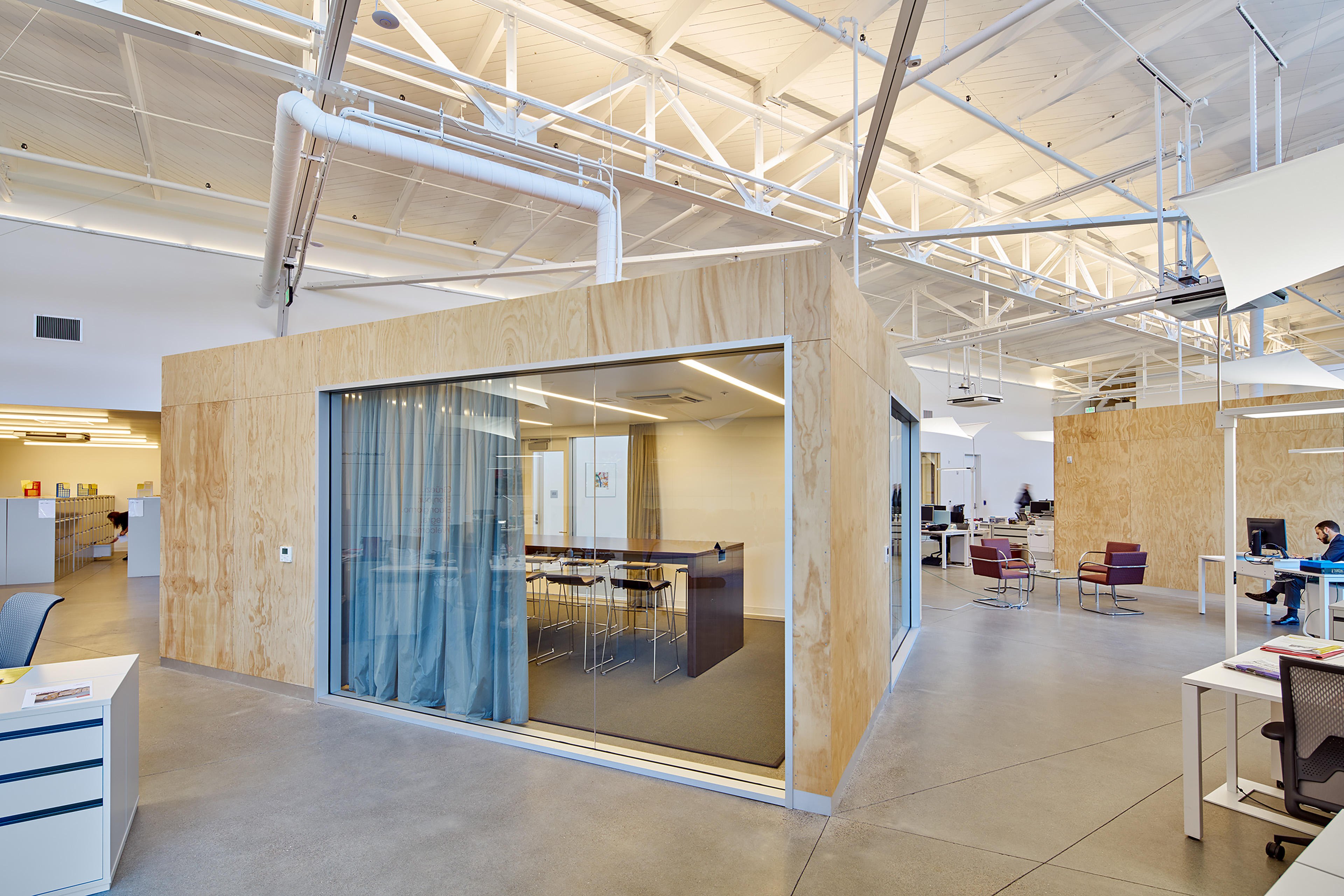 Open office with plywood-clad meeting rooms, glass-walled conference space, exposed white roof structure, desks, and lounge seating.