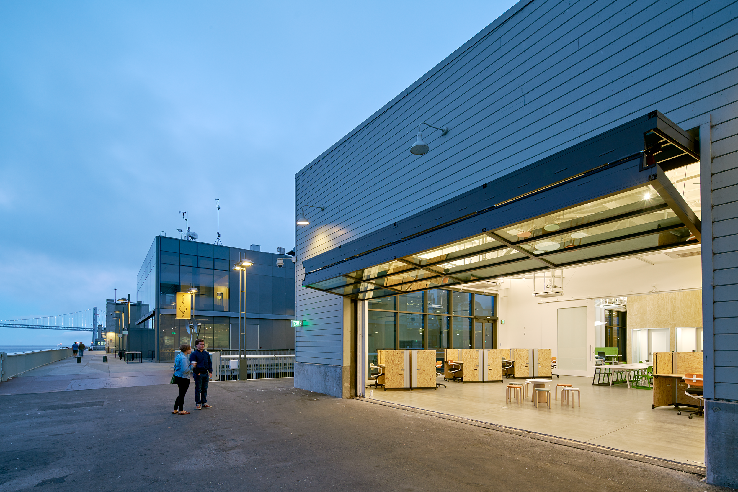 Exterior view of a waterfront office building at dusk with a large open garage-style door revealing a bright workspace, plywood desks, and seating inside.