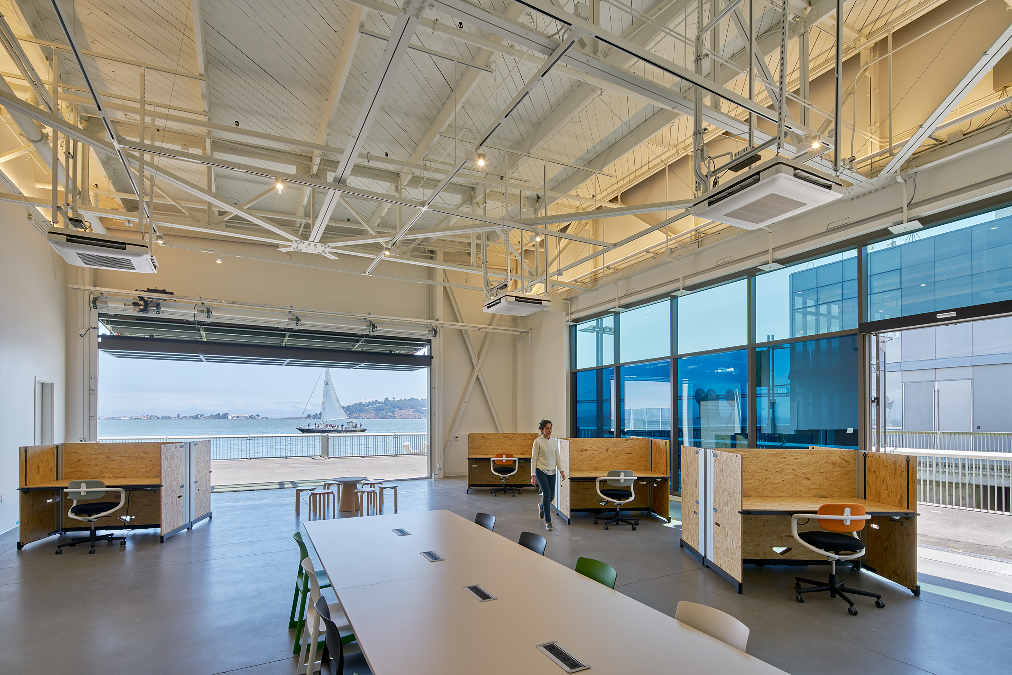 High-ceilinged waterfront workspace with exposed white trusses, plywood workstations, a large shared table, and an open garage door framing a view of the bay and a sailboat.