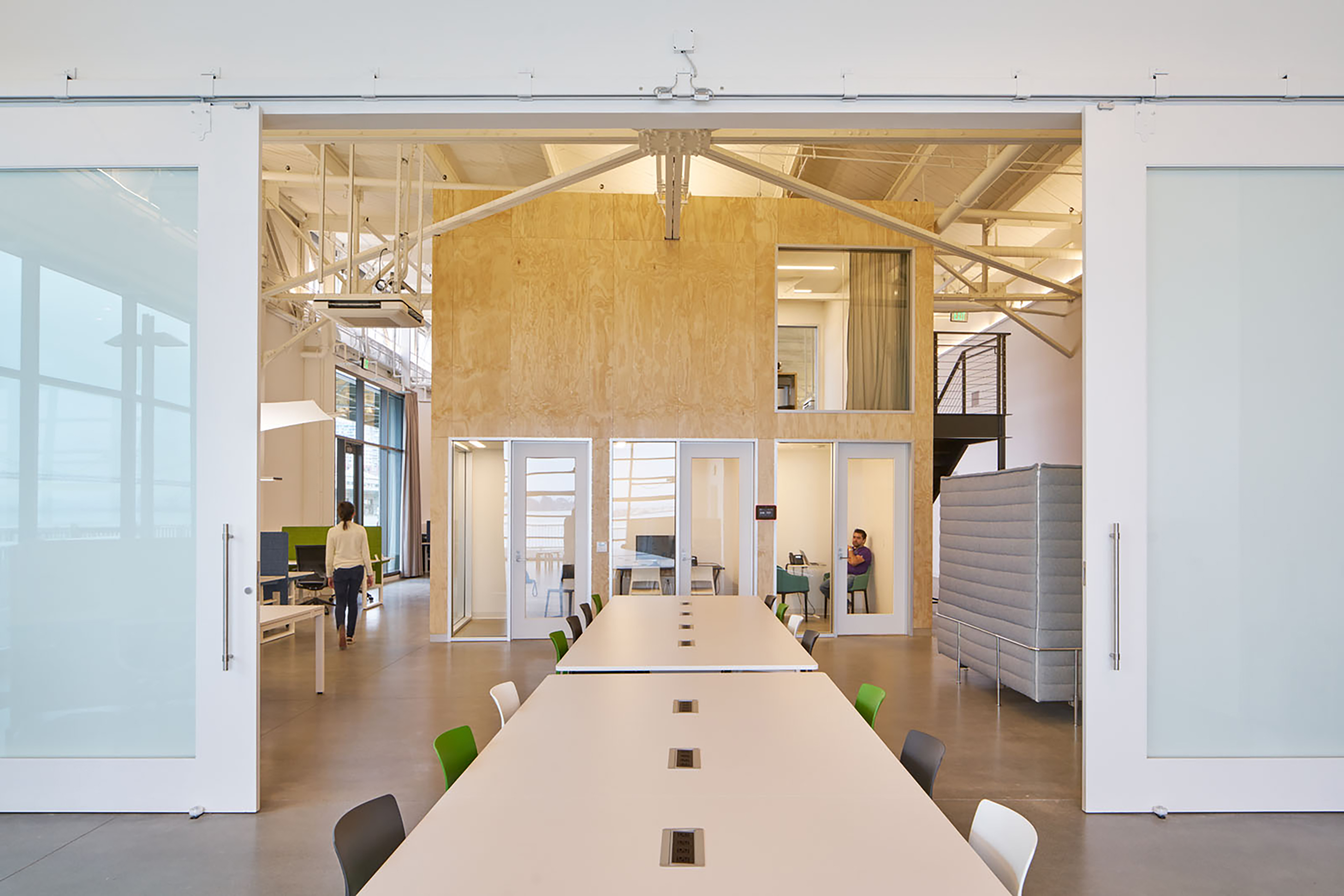 Long shared table leading into a bright open office with plywood-clad rooms, glass sliding doors, exposed white trusses, and people working in the background.