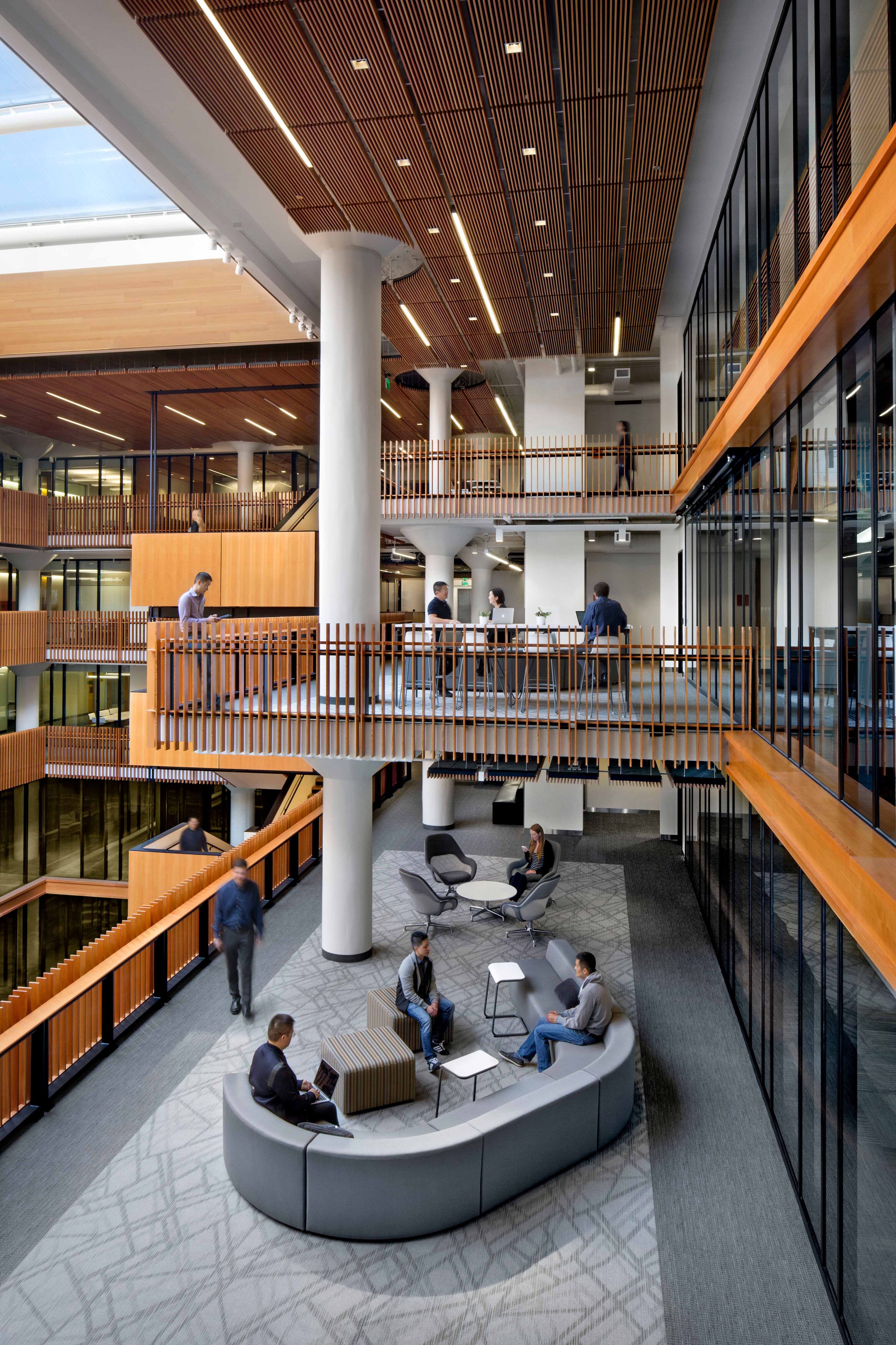 Multi-level office atrium with glass-fronted workspaces, wood railings and ceilings, open stairs, and lounge seating areas that support informal meetings and collaboration.