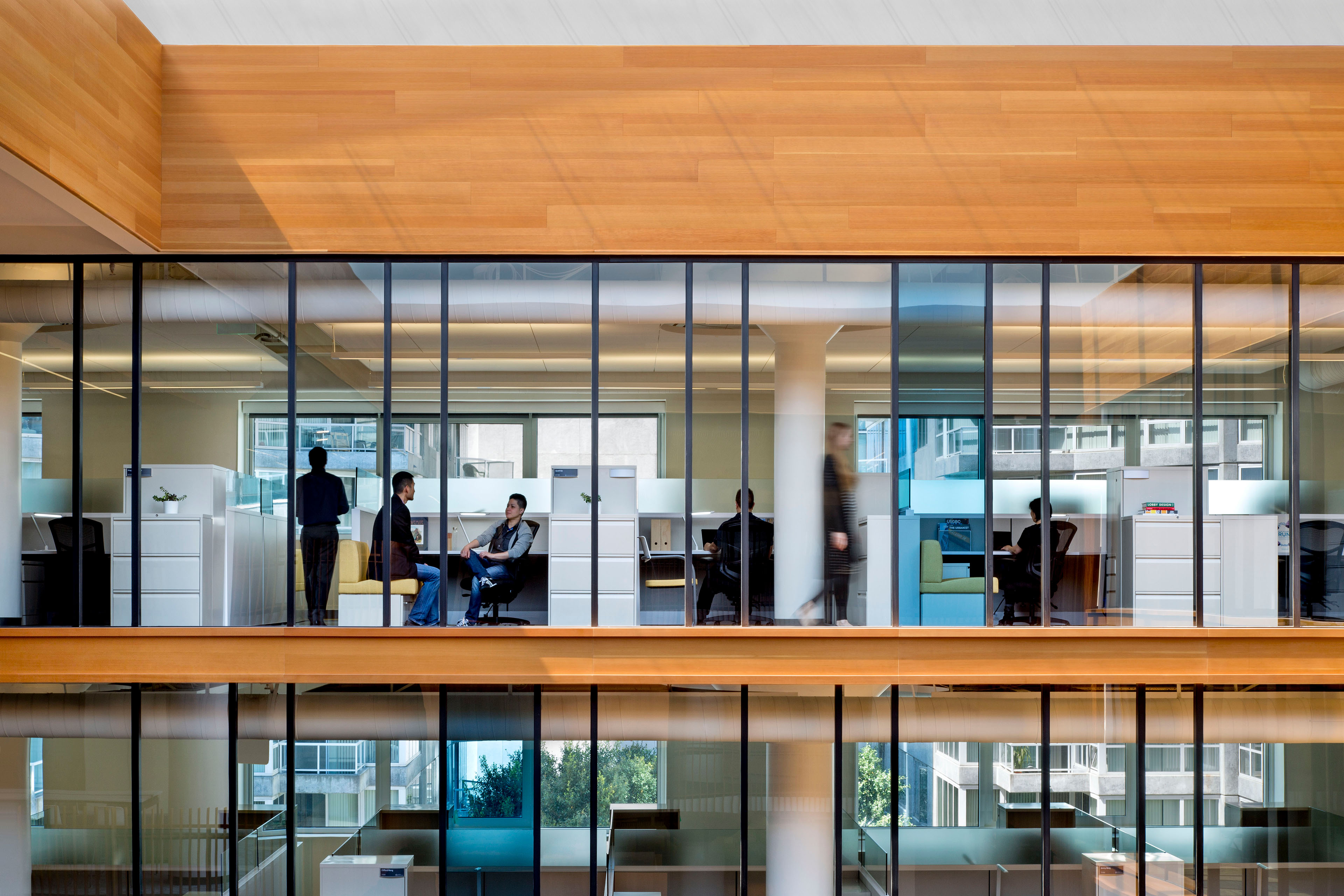 Glass-walled office interior showing individual workstations, informal seating, wood ceiling accents, and people working across multiple levels.