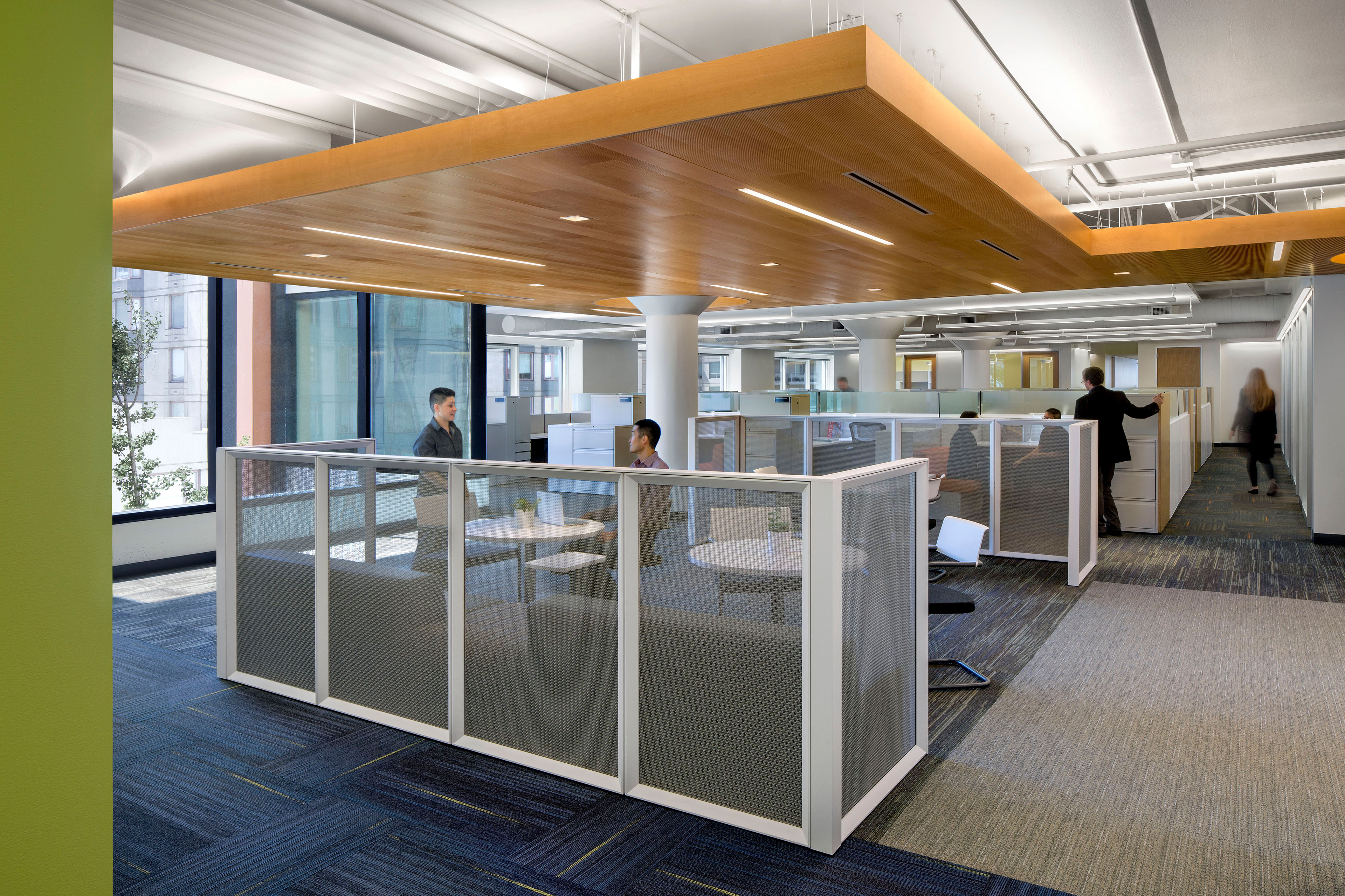 Open office workspace with low-partition collaboration zones, individual workstations, wood ceiling accents, and large windows bringing daylight into the interior.