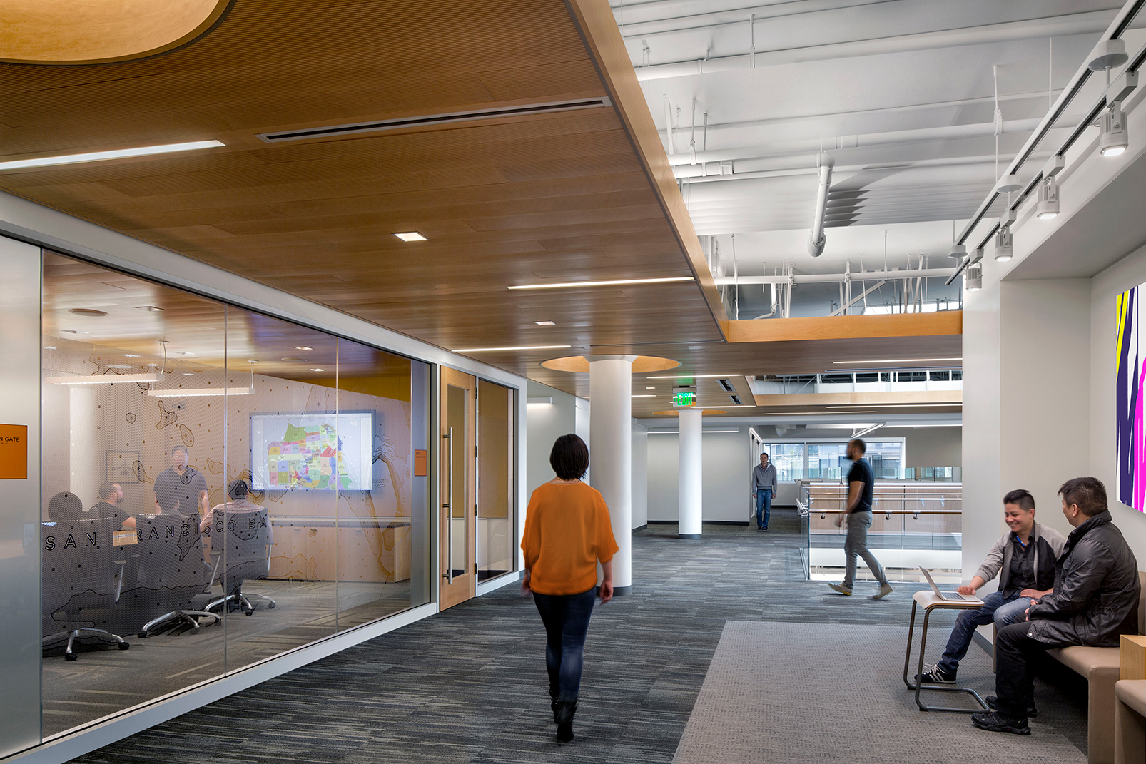 Office corridor with glass-walled meeting rooms, wood ceiling accents, bench seating, and people working and gathering near an open atrium.