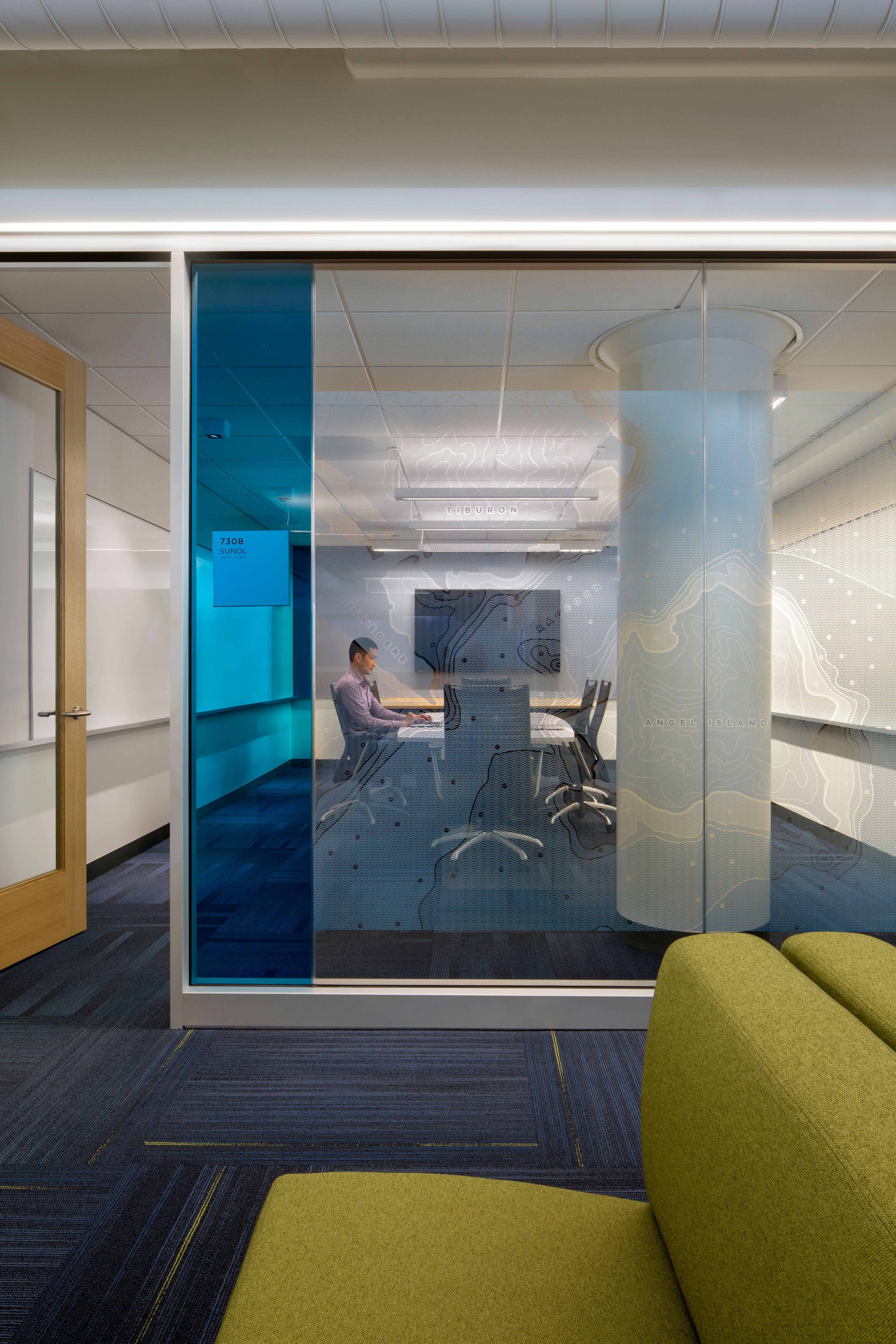 Glass-walled conference room with map graphics and blue film on the glazing, showing a person working at a meeting table inside and lounge seating in the foreground.