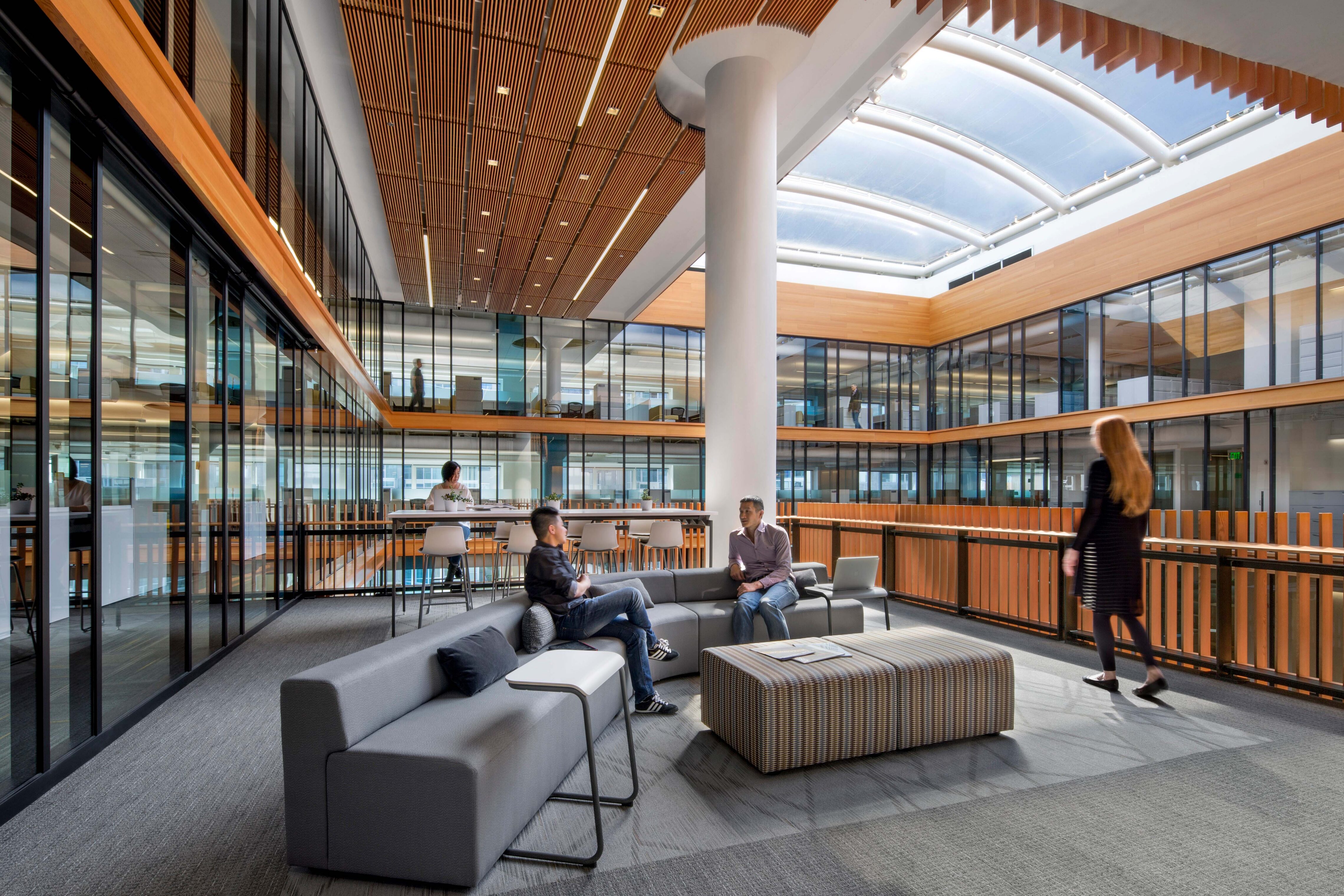 Multi-level office atrium with glass-fronted workspaces, wood railings and ceilings, lounge seating, and a large skylight overhead.