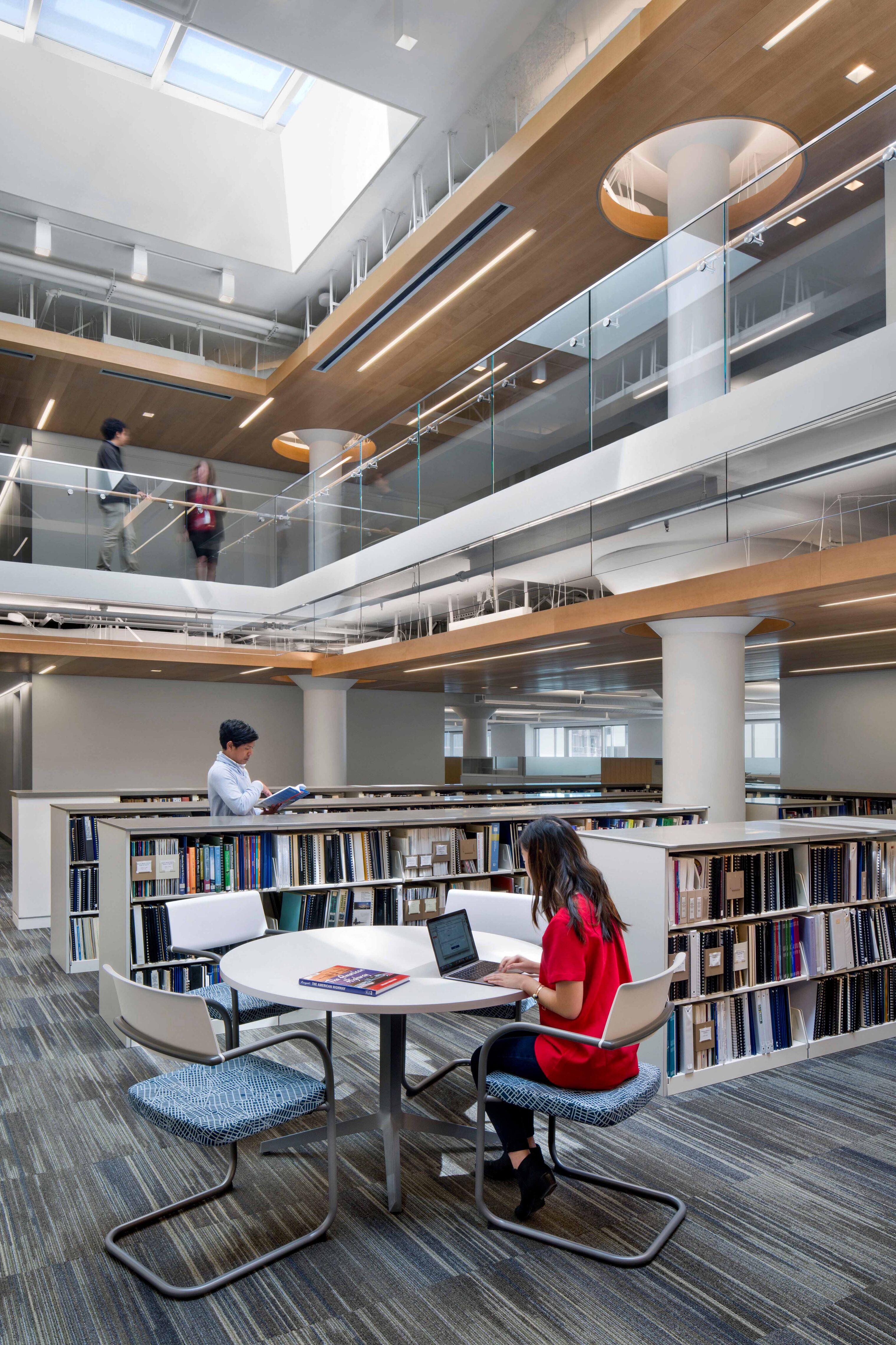 Multi-level workplace library with book stacks, round study tables, glass railings, wood ceiling accents, and skylights bringing daylight into the atrium.