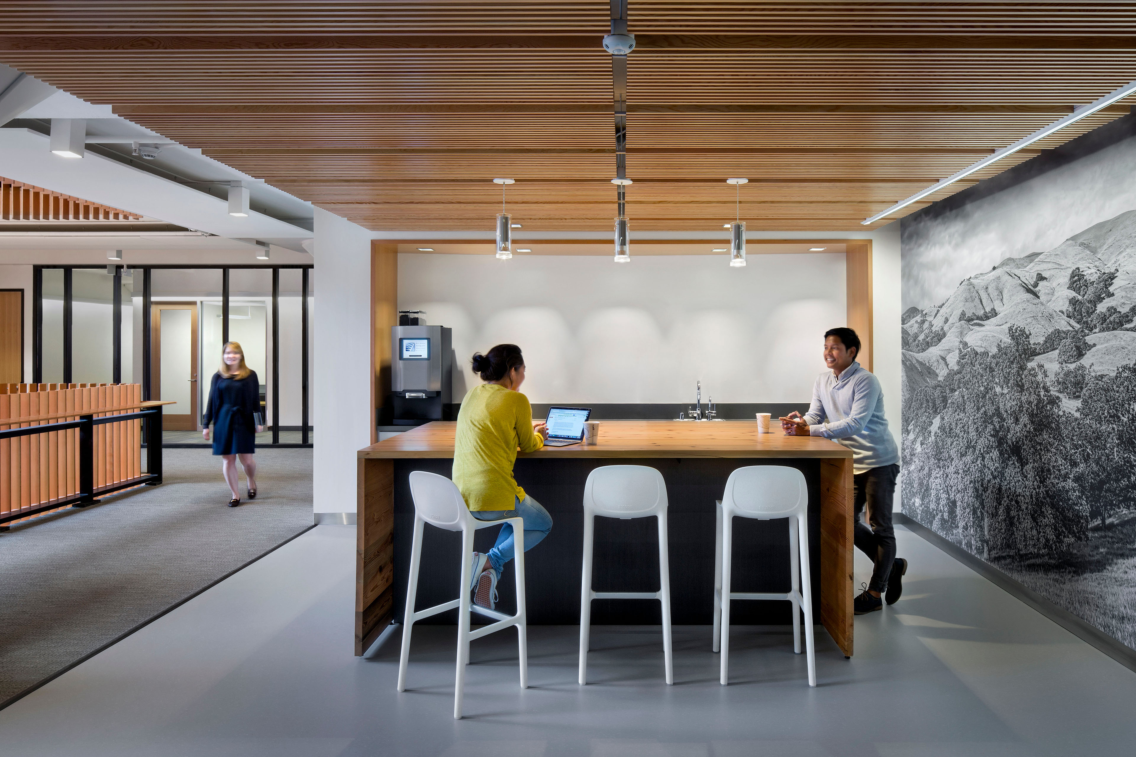 Office kitchen and gathering area with a wood counter, white stools, slatted wood ceiling, and a large landscape mural supporting informal collaboration.