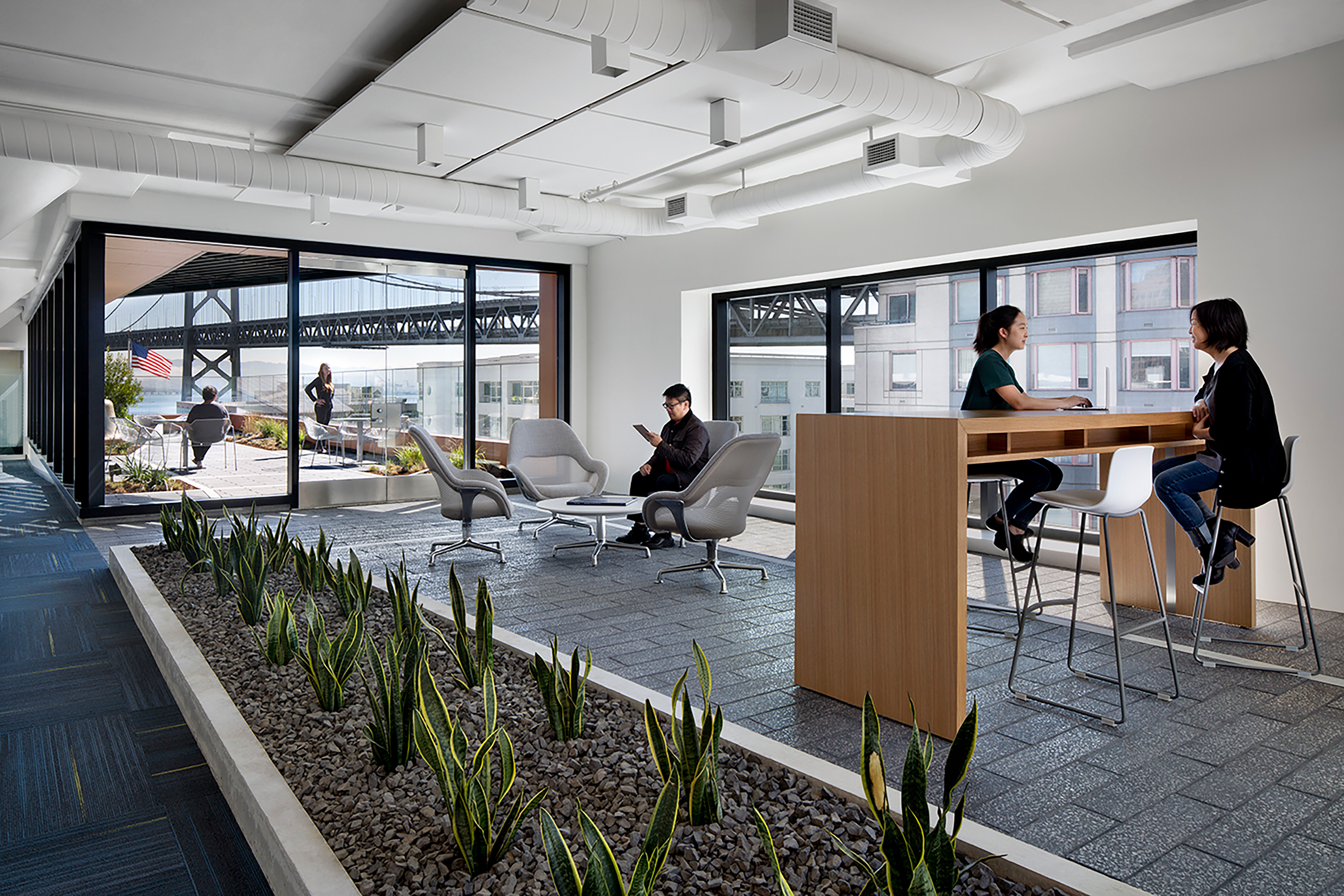 Interior office lounge with planted areas, soft seating, high tables, and direct access to an outdoor terrace with views of the Bay Bridge.