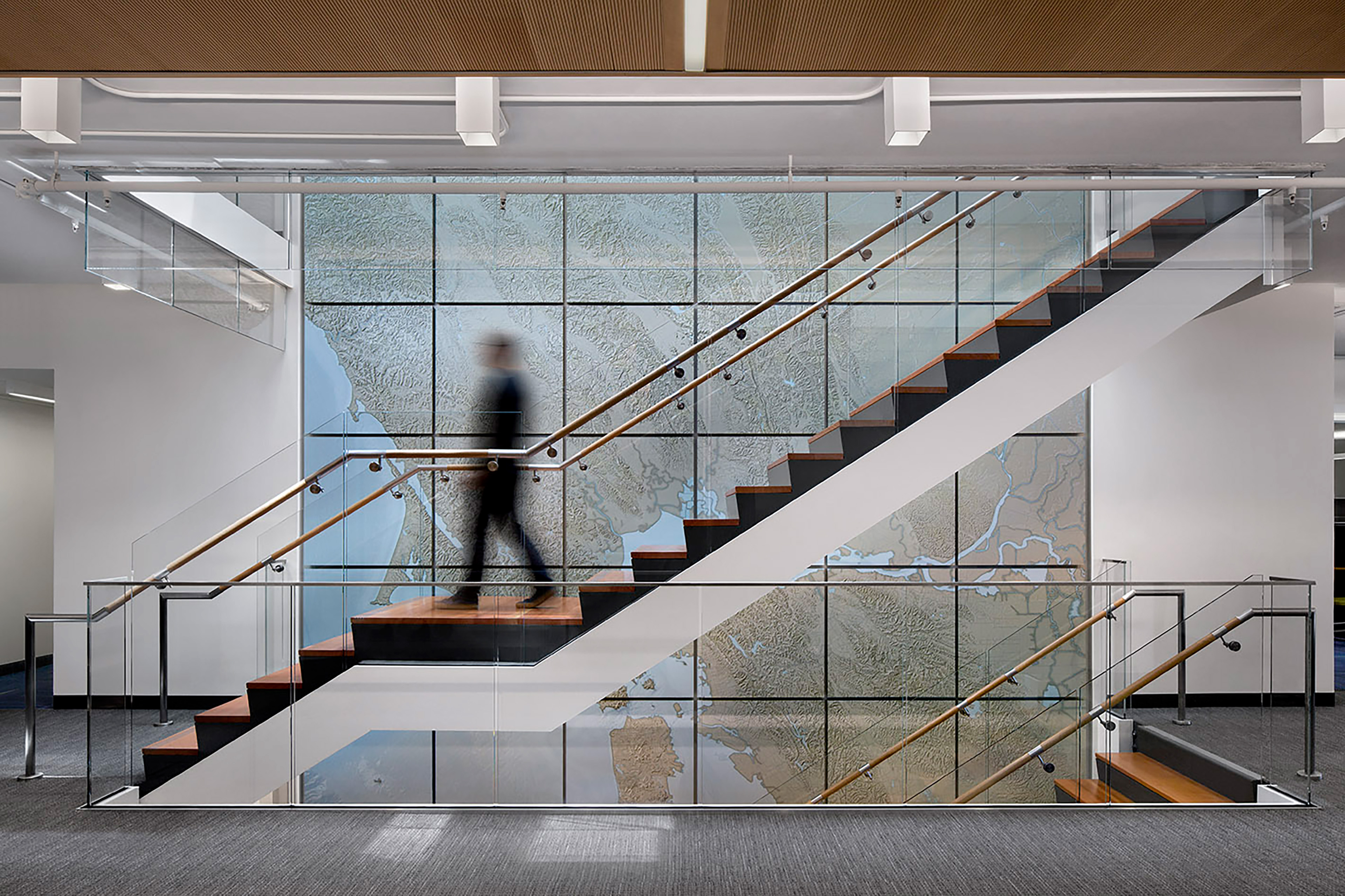 Interior stair with glass railings, wood treads, and a large map graphic wall, with a person moving through the multi-level office space.