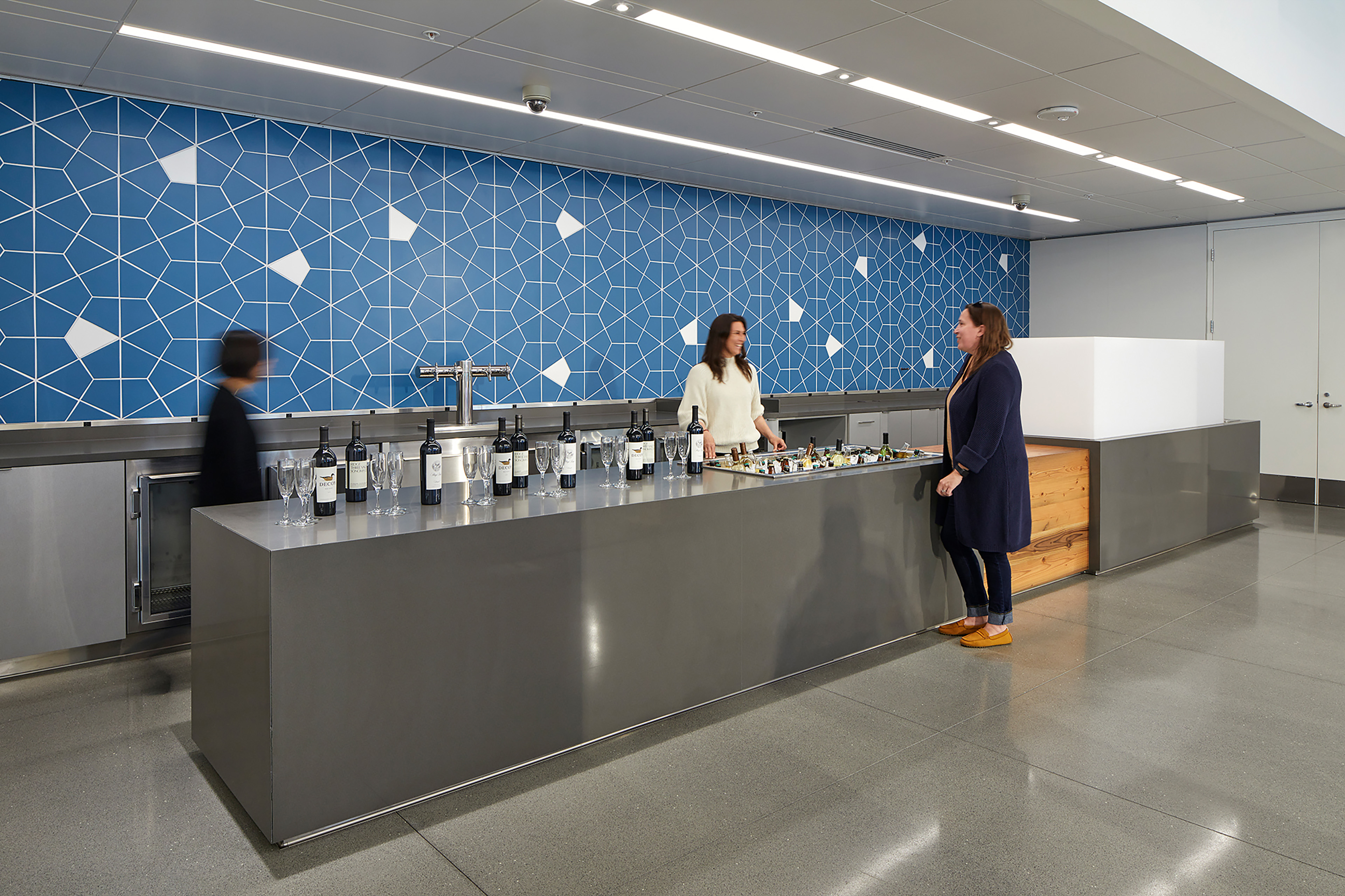 Event bar area with a stainless steel counter, blue geometric tile wall, wine bottles, and people gathering in a flexible workplace hospitality space.