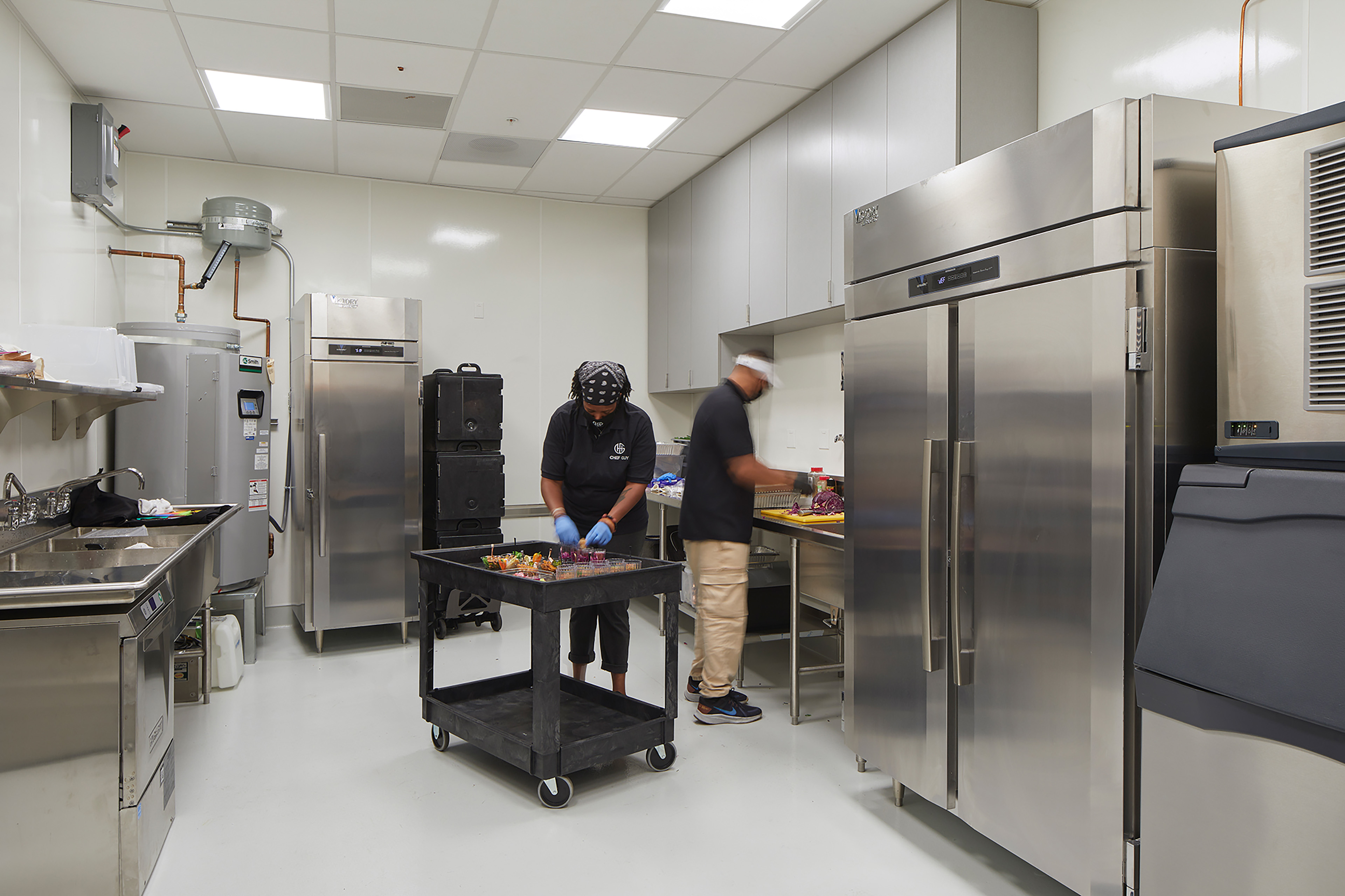 Commercial kitchen with stainless steel appliances, refrigerators, prep tables, and staff preparing food in a back-of-house workspace.