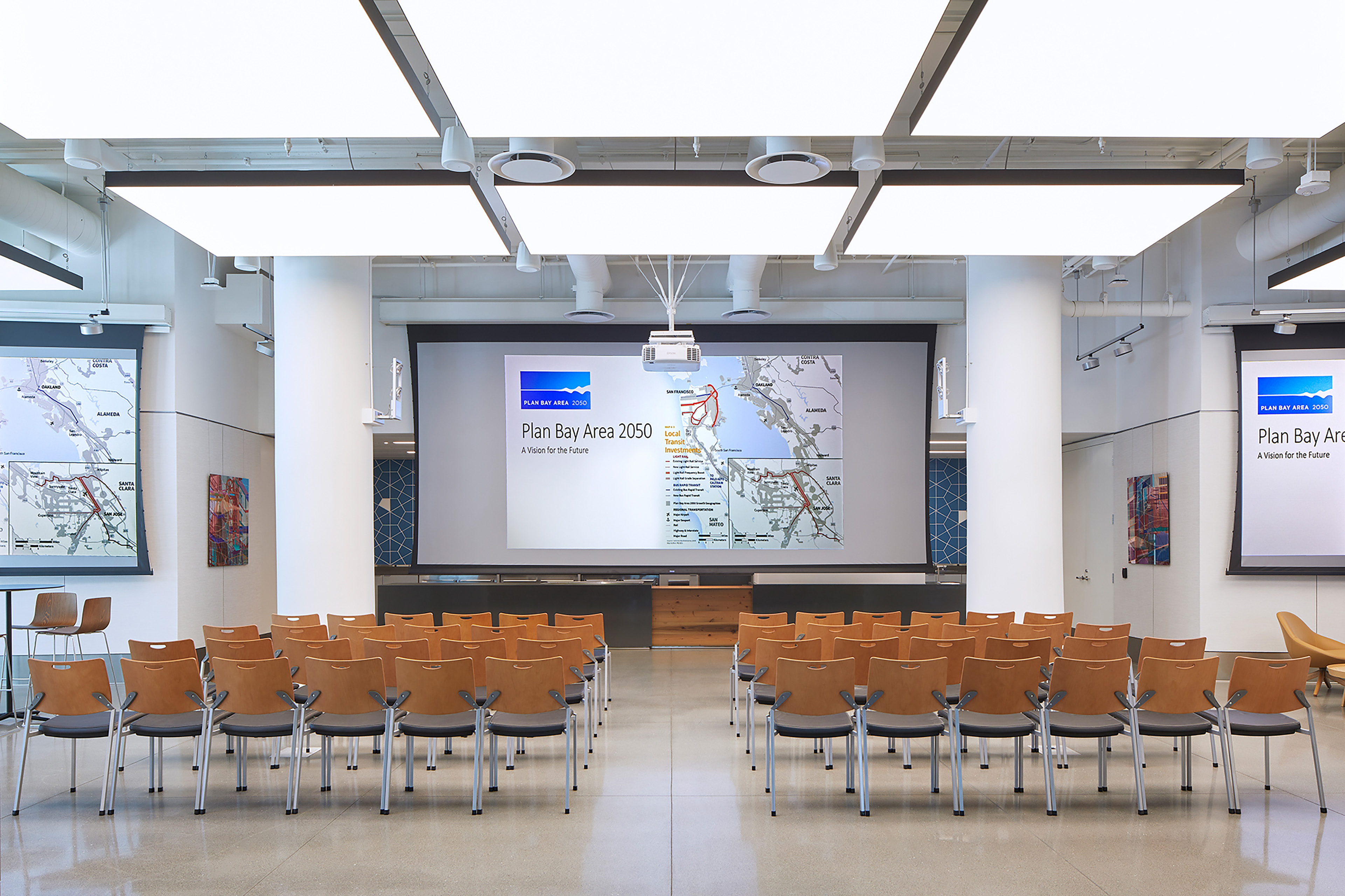 Flexible presentation room arranged in lecture-style seating. Rows of wood-backed chairs face a large central projection screen displaying a “Plan Bay Area 2050” presentation, with matching content shown on two side screens. Large luminous ceiling panels and exposed white mechanical systems brighten the open interior.