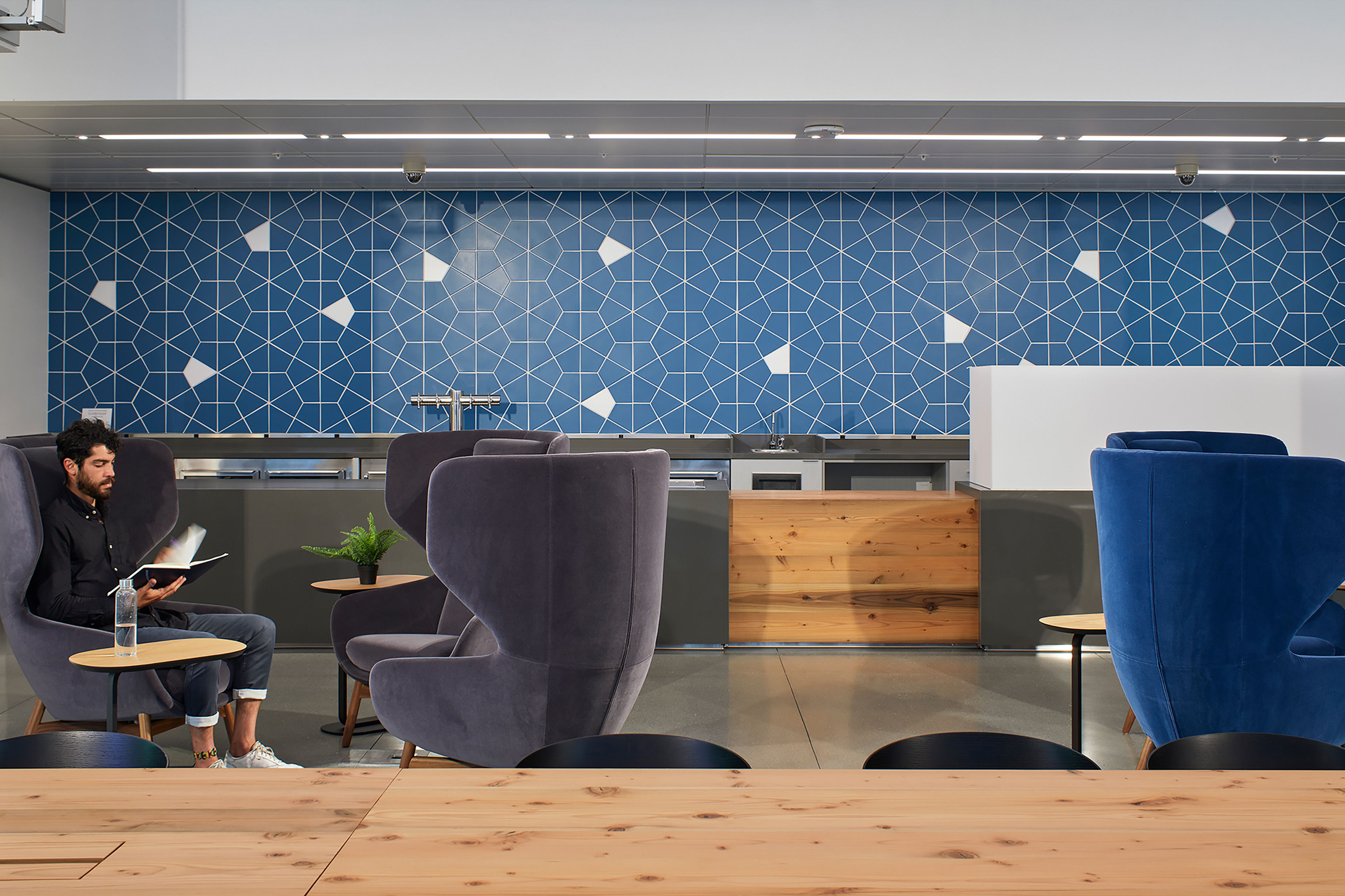 Lounge and café area with high-back chairs, small tables, and a long wood communal table. A person reads in one of the chairs, with a blue geometric tile wall and kitchenette counter in the background.
