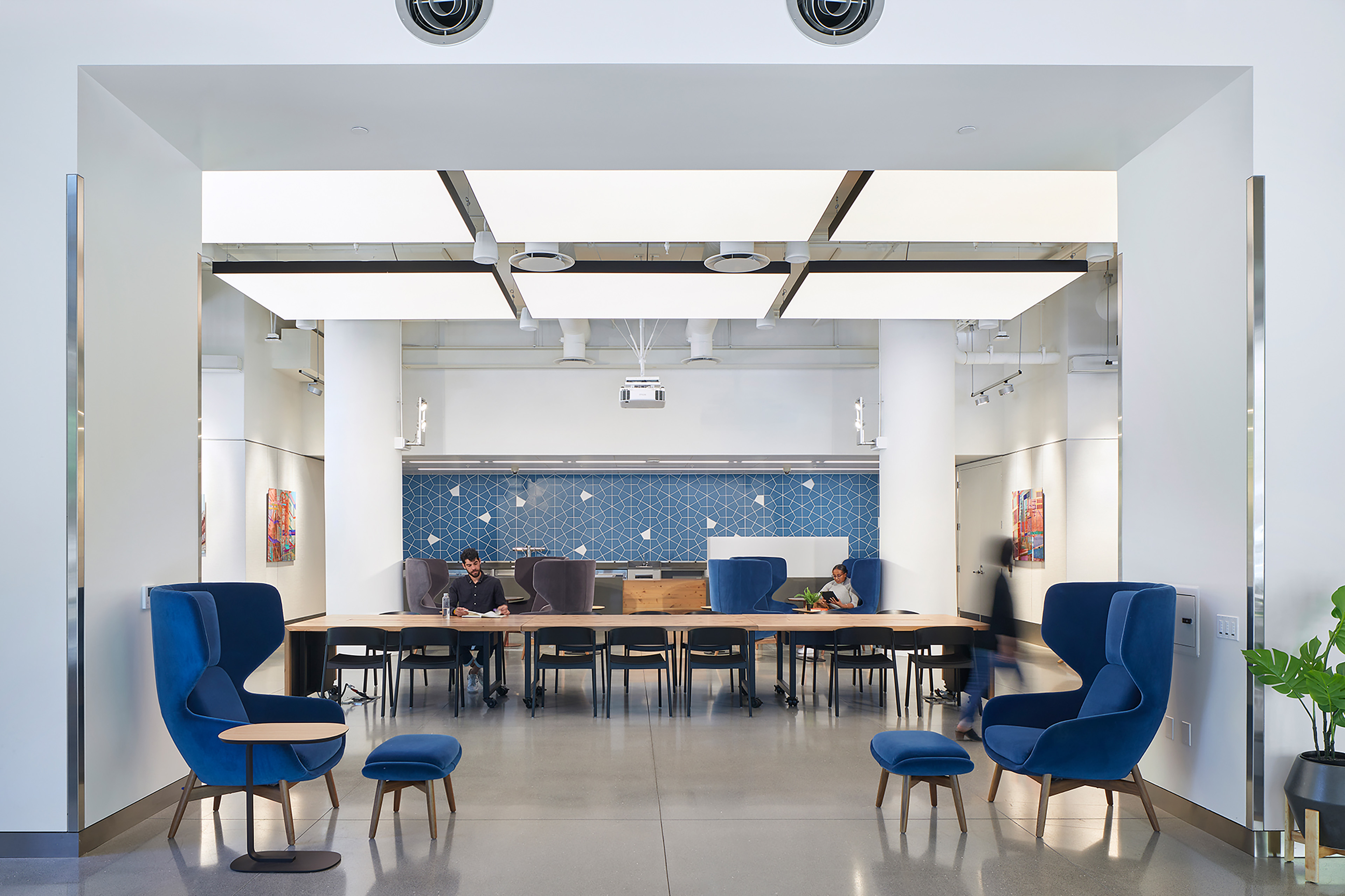 Bright, modern collaboration space framed by a wide white opening. Two blue lounge chairs with matching ottomans sit in the foreground, while a long communal table with black chairs extends across the center. A few people work in the background beneath large illuminated ceiling panels, with a blue geometric tile wall behind them.