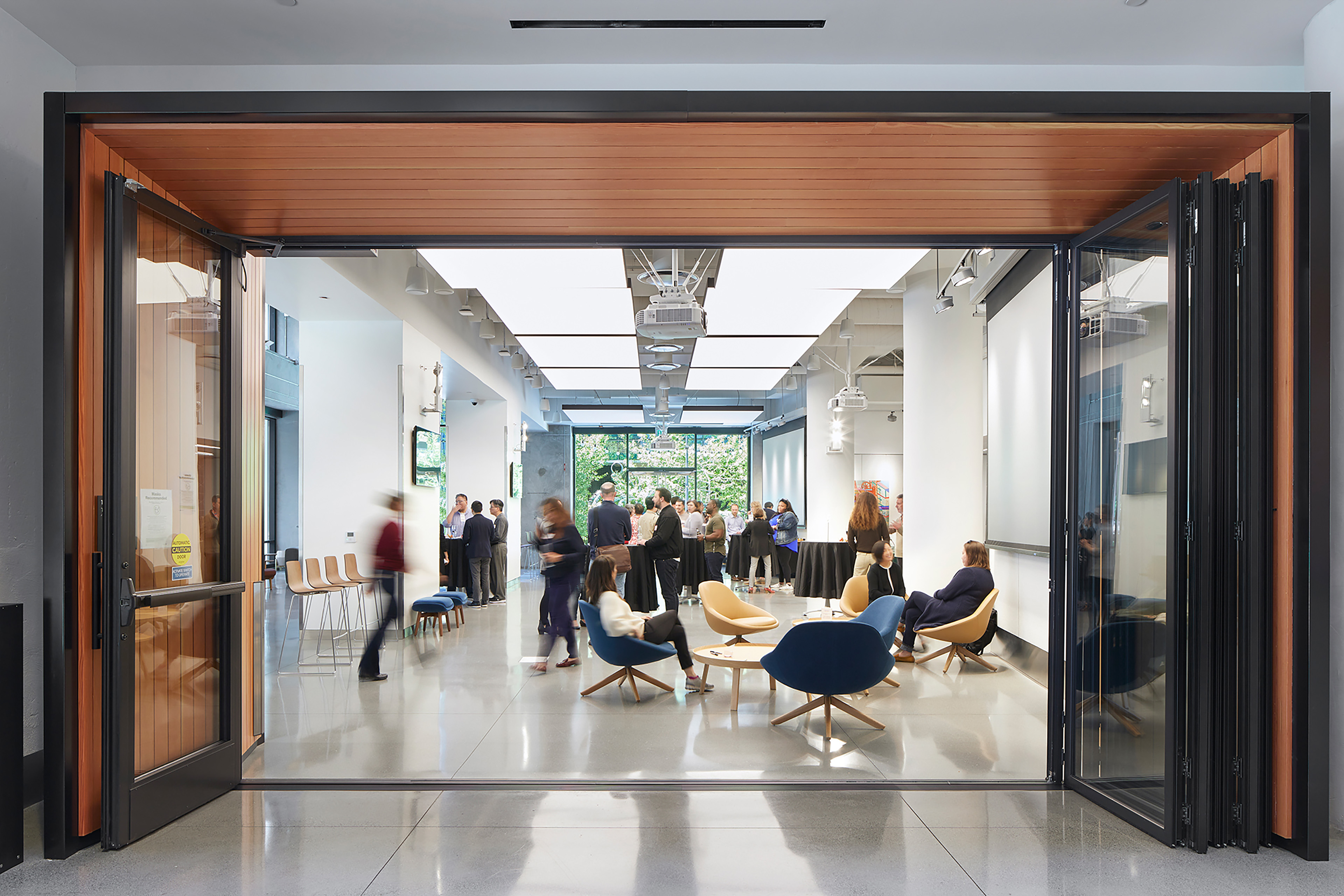 Flexible event space viewed through large open folding glass doors, with a wood-lined entry frame, polished concrete floor, illuminated acoustic ceiling panels, lounge seating in the foreground, and people gathering near café tables and presentation screens in the background.