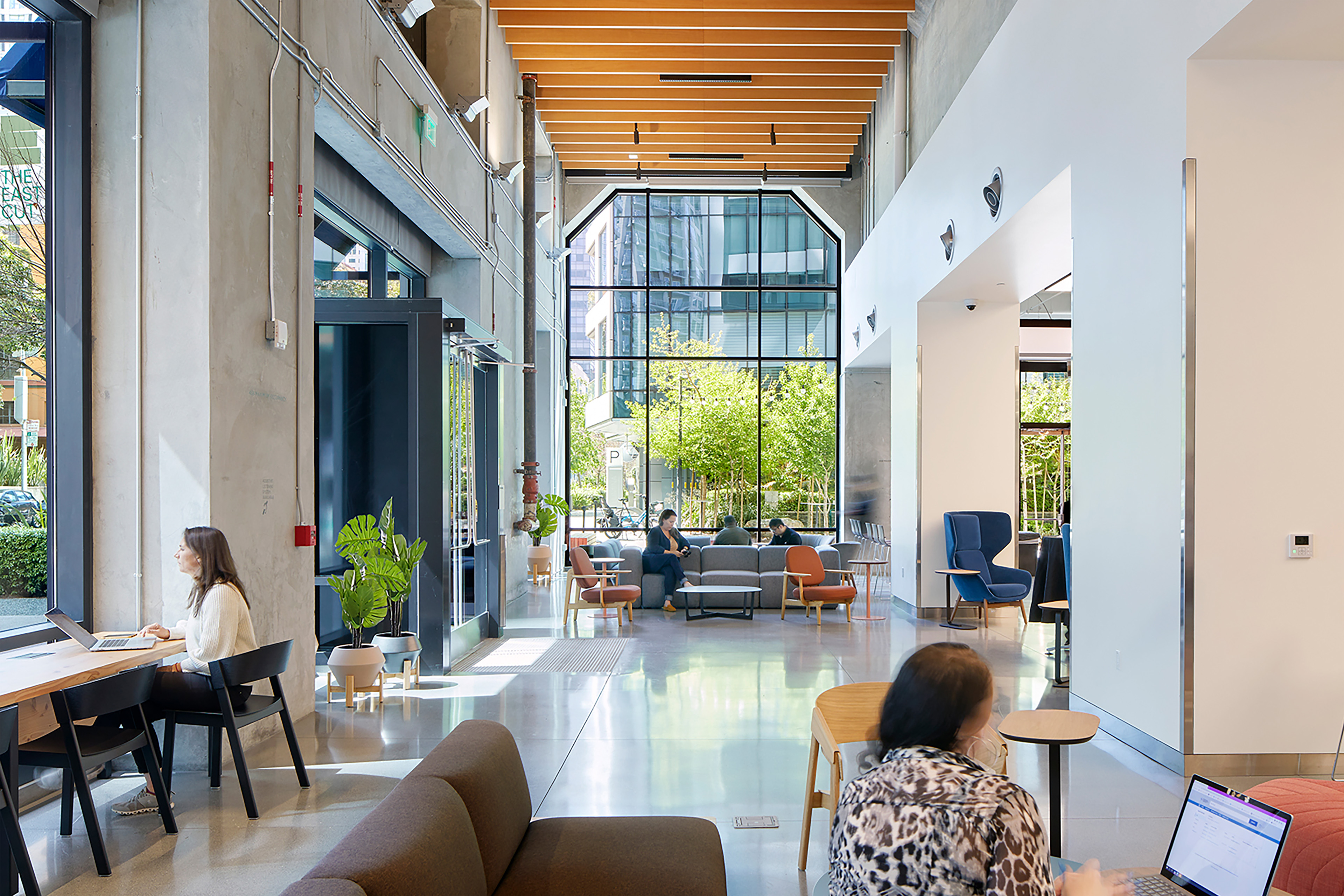 Bright workplace lobby with exposed concrete walls, wood ceiling accents, polished floors, lounge seating, and people working near large windows overlooking the street.