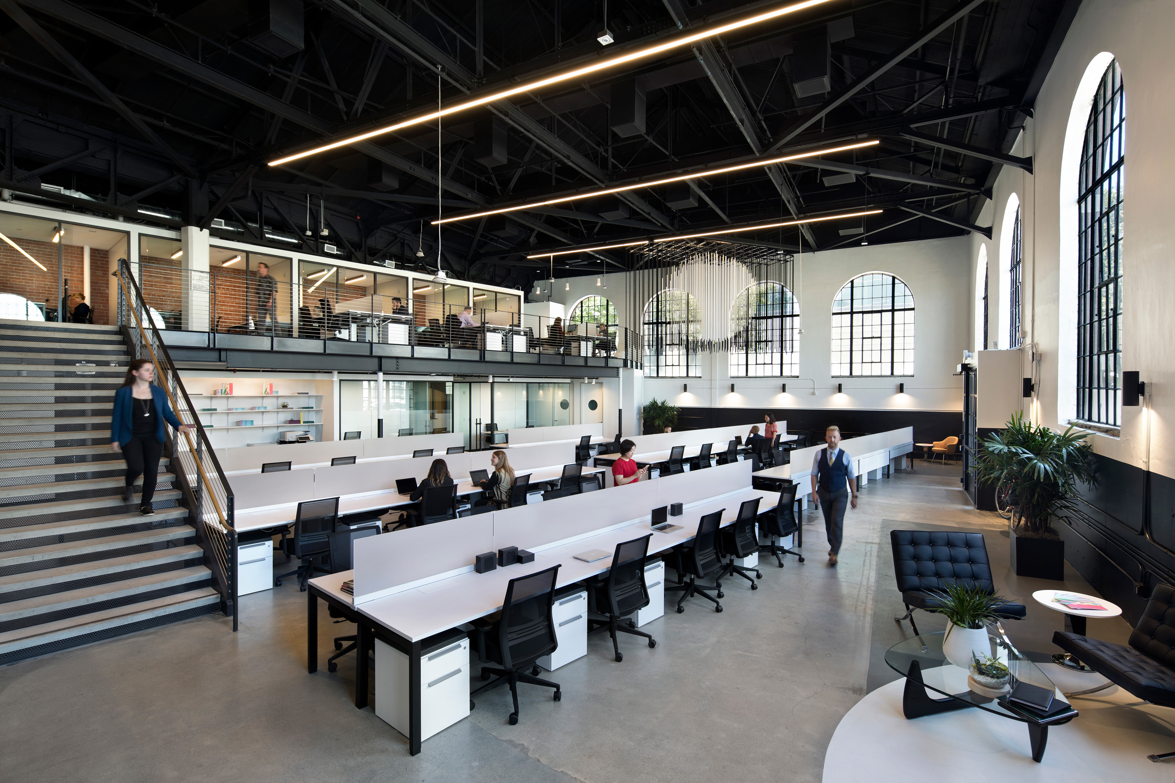 Open workplace in a historic building with high ceilings, arched windows, exposed black trusses, and long shared workstations. A mezzanine with glass-fronted offices overlooks the main work area, where employees work and move through the light-filled space.