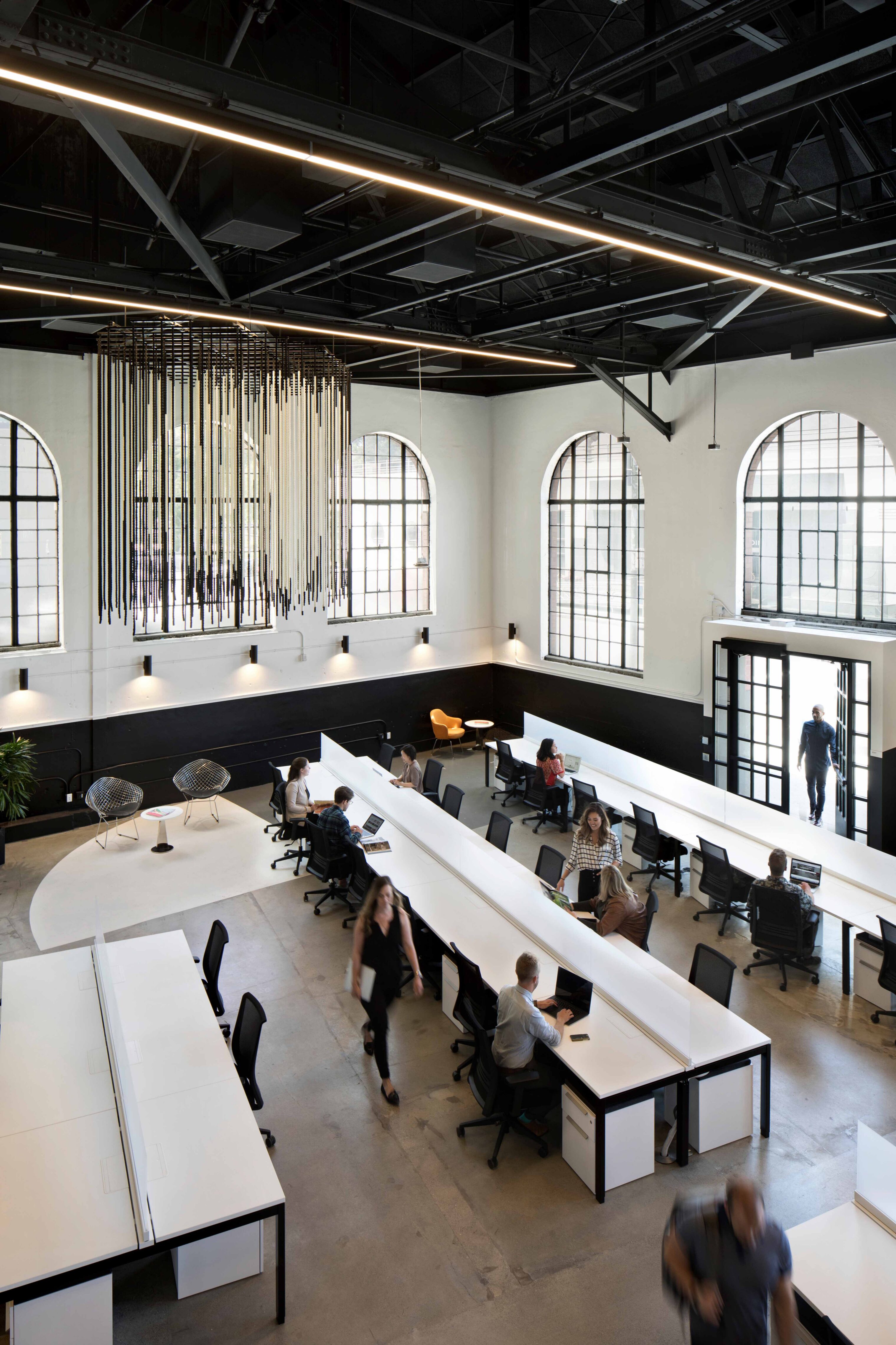Open workplace in a historic building with tall arched windows, exposed black structure, and long shared workstations. Employees work and move through the light-filled space below a large suspended decorative installation.