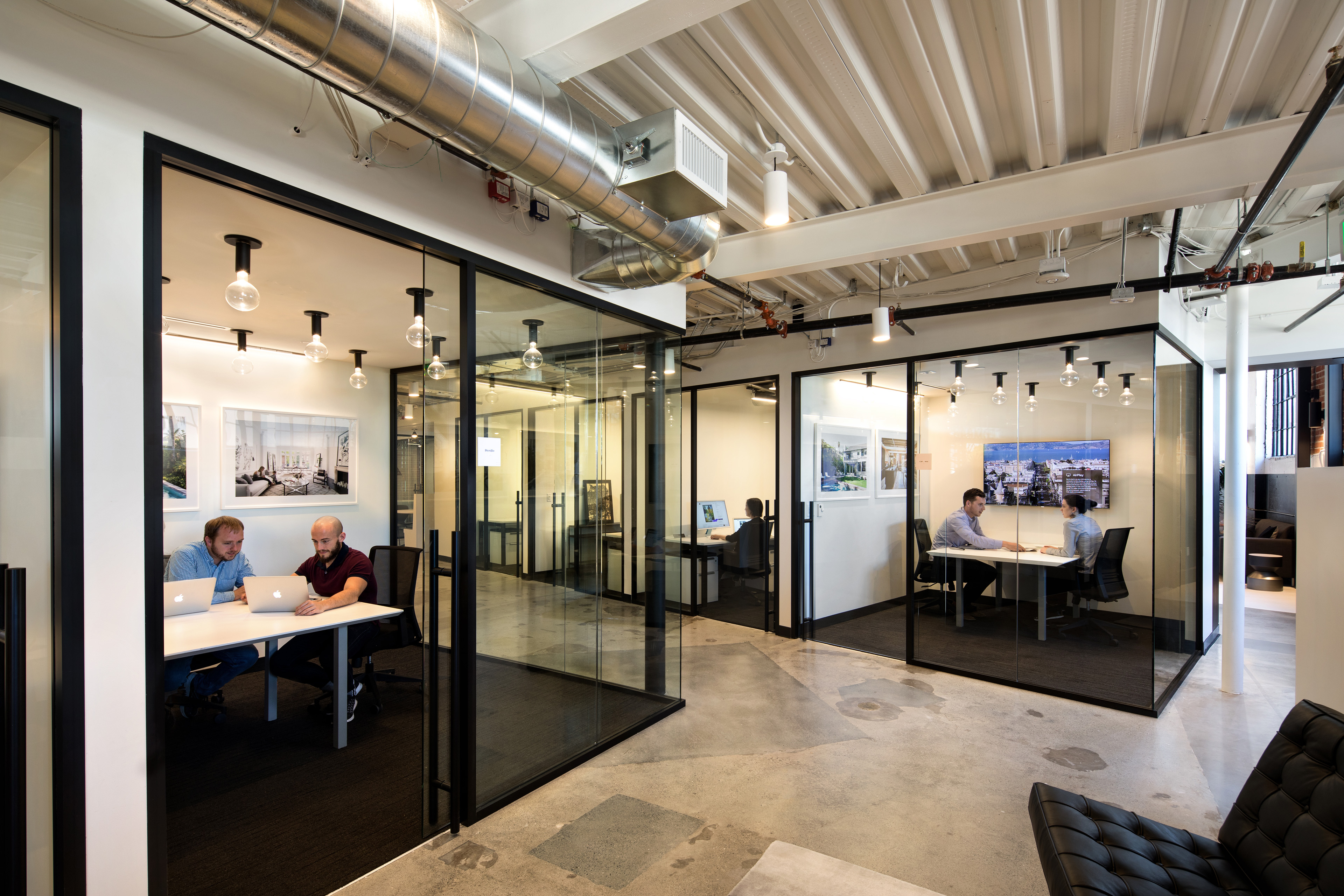 Glass-enclosed meeting rooms and offices in a modern workplace with exposed ductwork, concrete floors, and black-framed partitions. Small teams meet at desks inside the rooms, with project imagery and a wall-mounted display visible in the background.