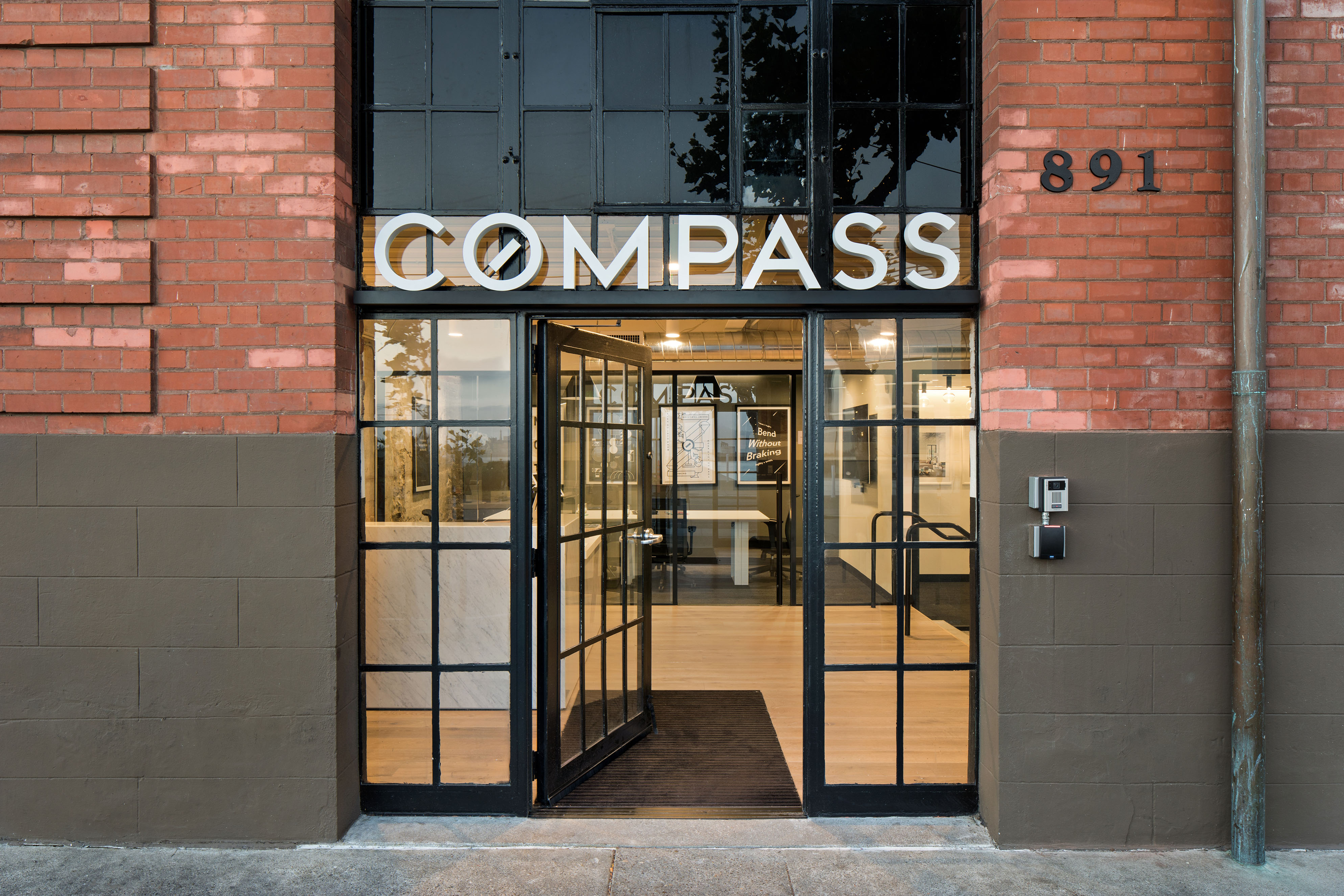 Close-up of the Compass entrance set into a historic brick facade, with black-framed glass doors and large white signage above. The open doorway reveals a bright interior with wood flooring, desks, and reception space beyond.