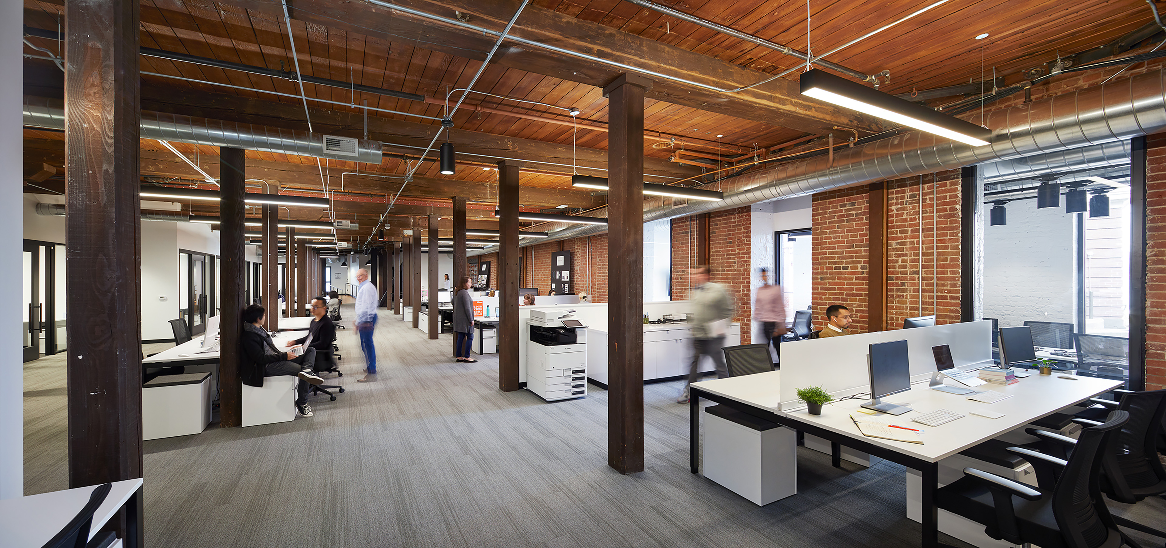 Open workplace in a historic brick-and-timber building, with exposed columns, wood ceilings, visible ductwork, and long shared workstations. Employees work and move through the space, where contemporary office furnishings sit within the preserved historic shell.