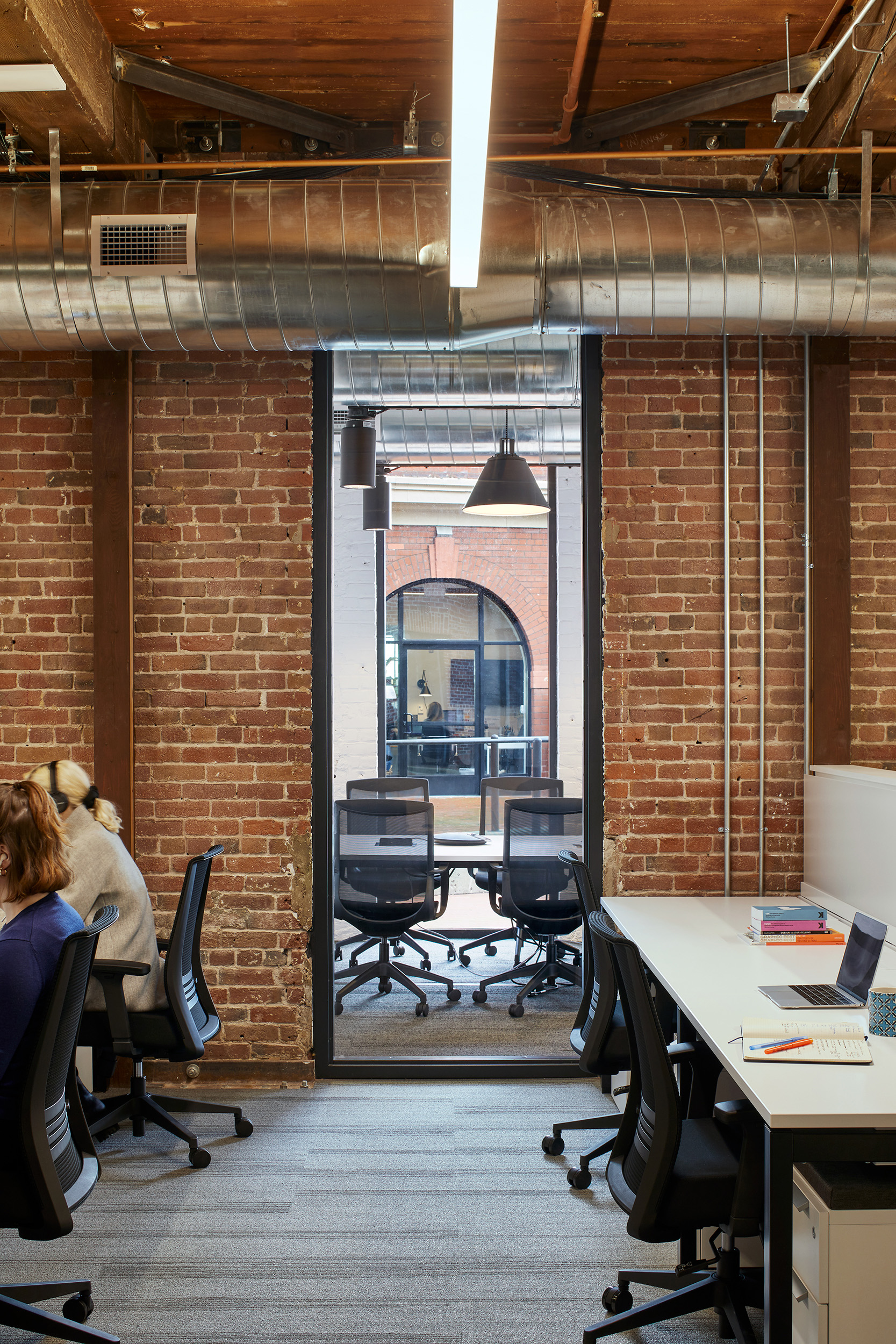 Open office with exposed brick walls, wood structure, visible ductwork, and glass-fronted meeting rooms. Workstations are arranged around the existing building fabric, with a view through to another conference space beyond.