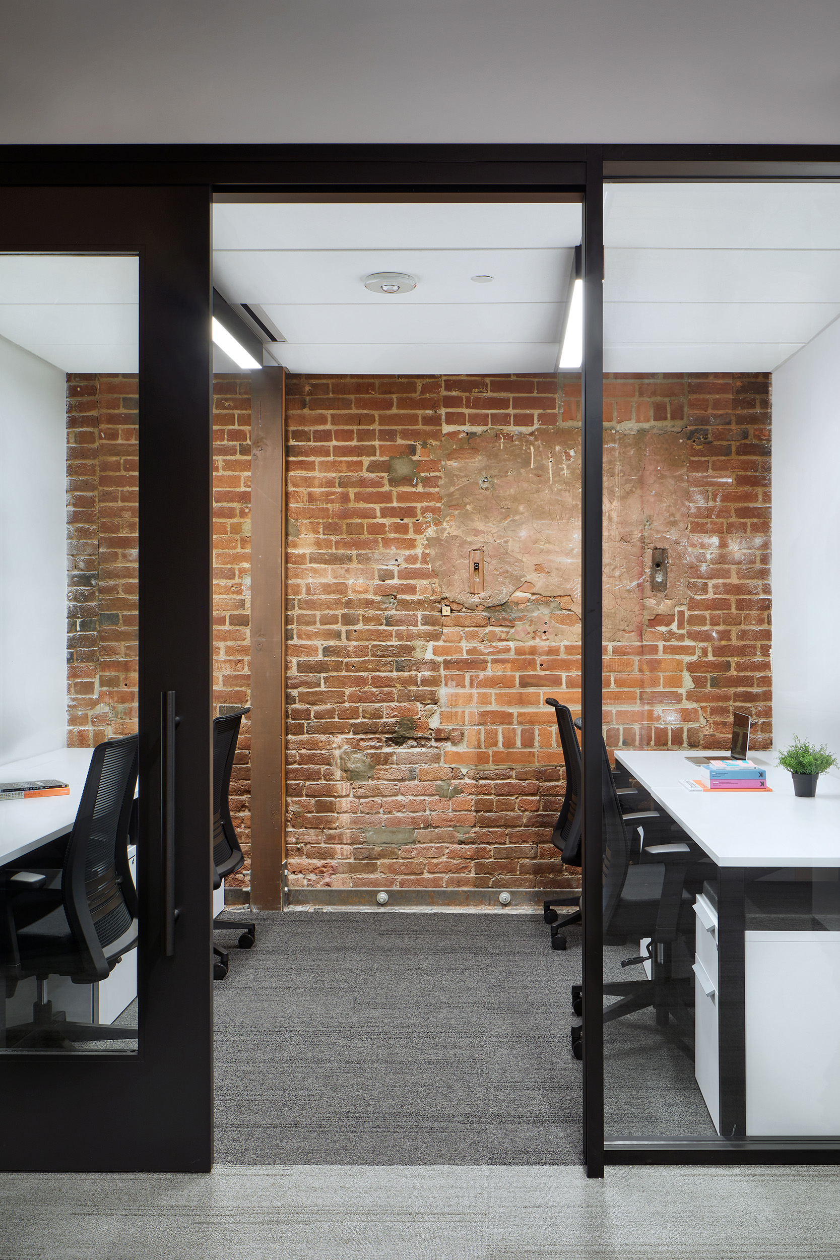 Glass-fronted office with exposed brick walls, modern workstations, and black-framed partitions. The historic masonry is left visible inside the room, contrasting with white desks, task chairs, and clean contemporary finishes.