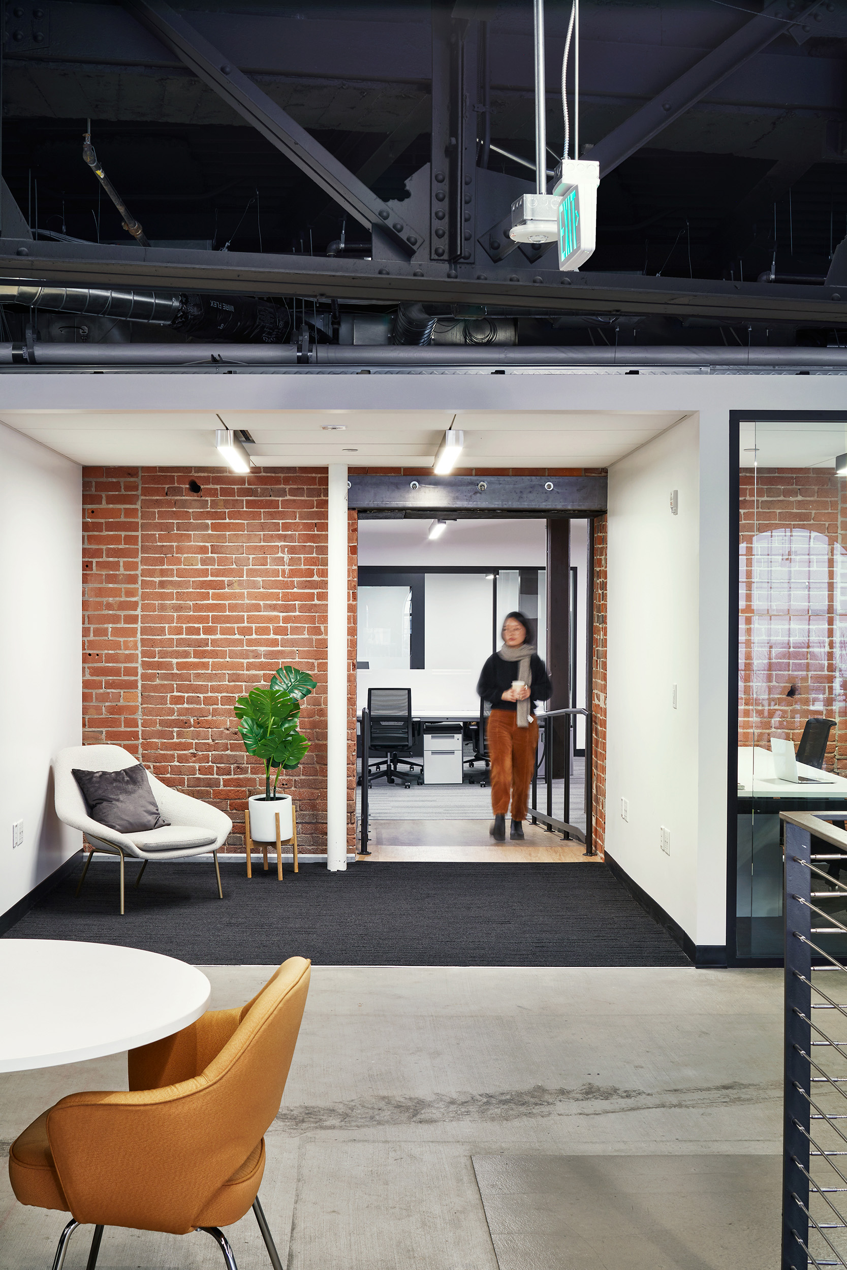 Lounge and transition area in an adaptive-reuse workplace, with exposed brick, steel structure, visible systems, and a small seating area. A person walks through the opening between work areas, framed by a historic masonry wall and contemporary office partitions.