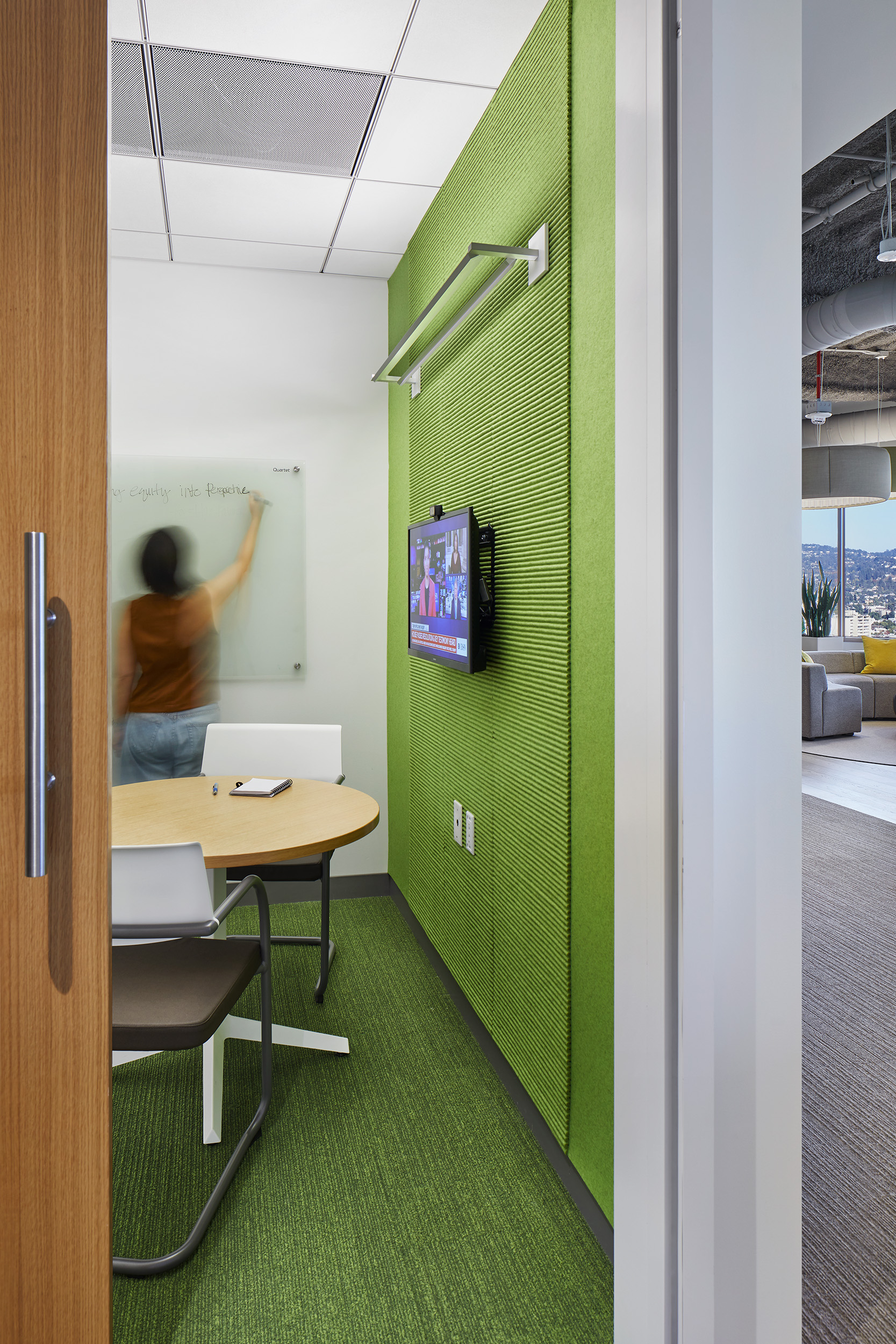 Small meeting room with a bright green acoustic wall, matching green carpet, round table, white chairs, wall-mounted monitor, and glass writing board. A person writes on the board, with the adjacent open lounge visible through the doorway.