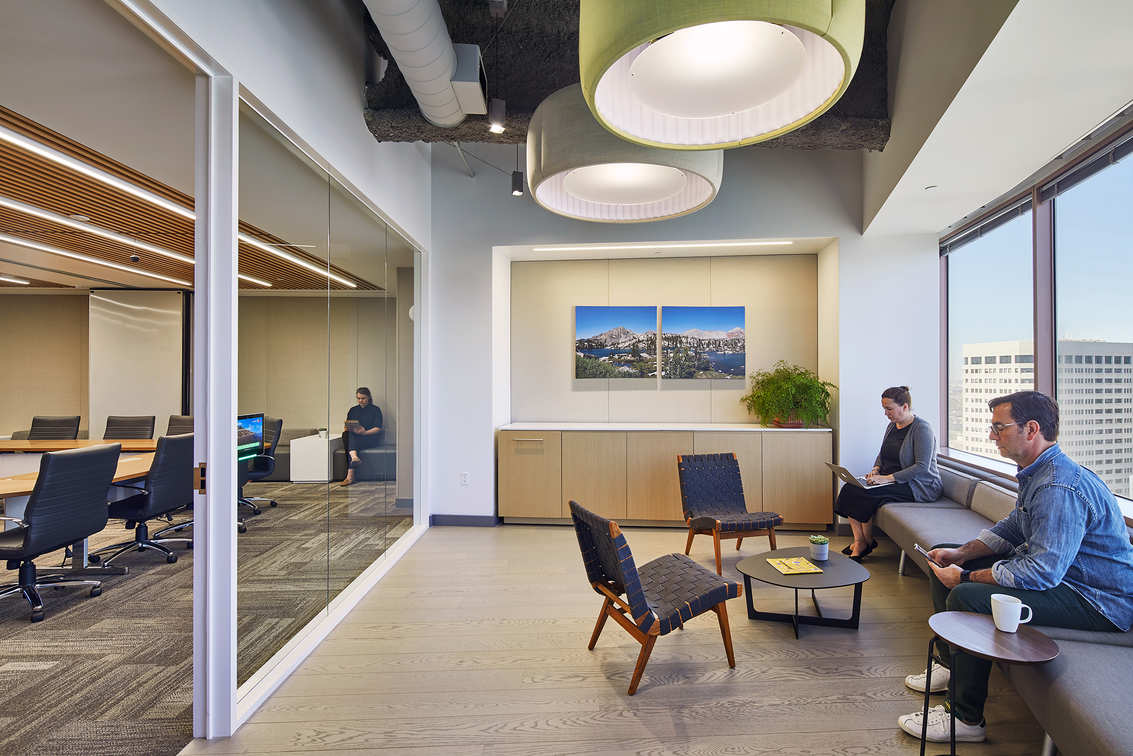 Lounge area beside a glass-enclosed conference room, with large circular pendant lights, light wood flooring, built-in cabinetry, low tables, and upholstered seating near expansive windows. Two people sit on a sofa working with devices, while another person sits inside the adjacent meeting room.