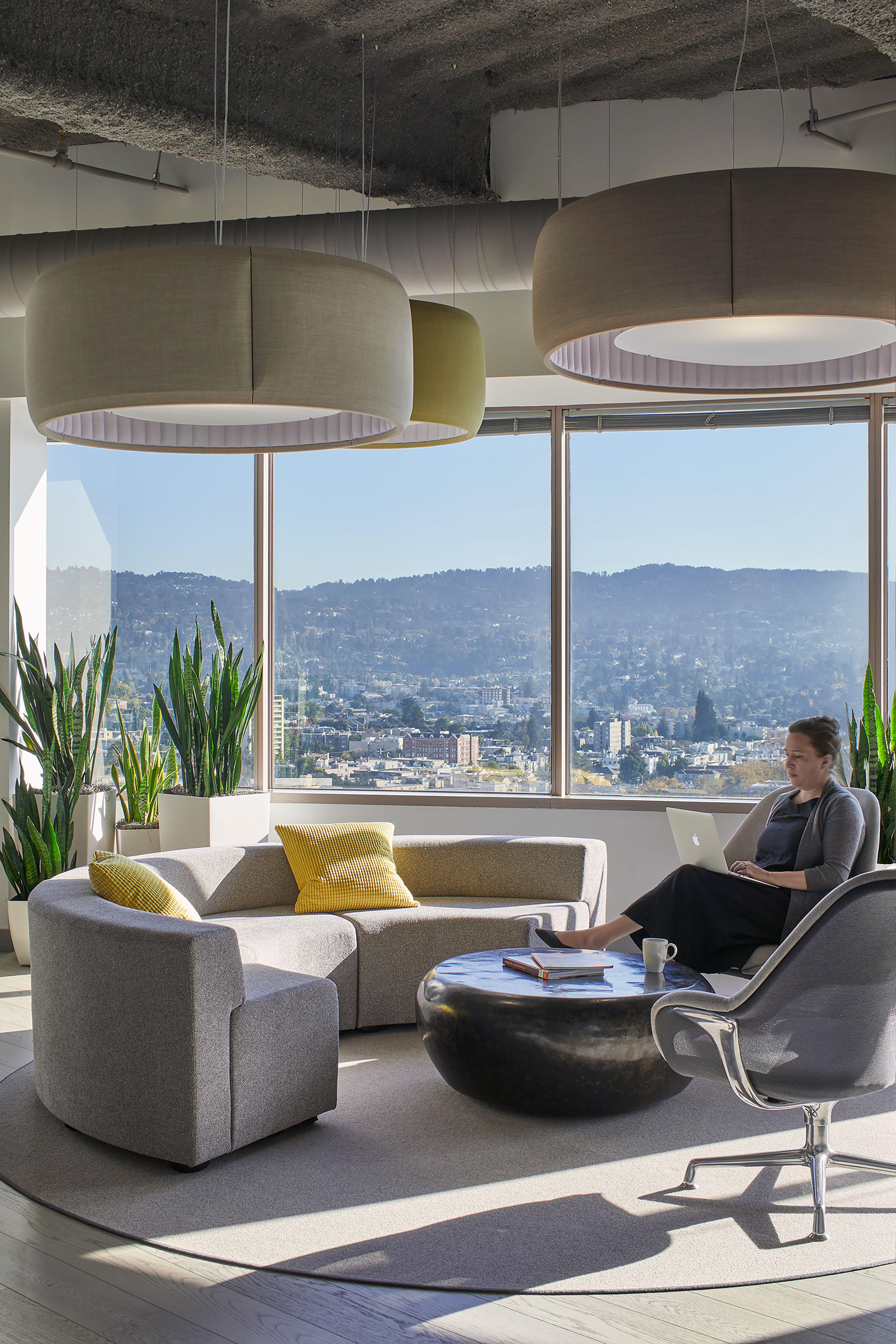 Bright office lounge with gray modular seating, yellow accent pillows, large circular acoustic pendant lights, tall indoor plants, and expansive windows framing a city and hillside view. A person sits in a gray lounge chair working on a laptop beside a low round coffee table.
