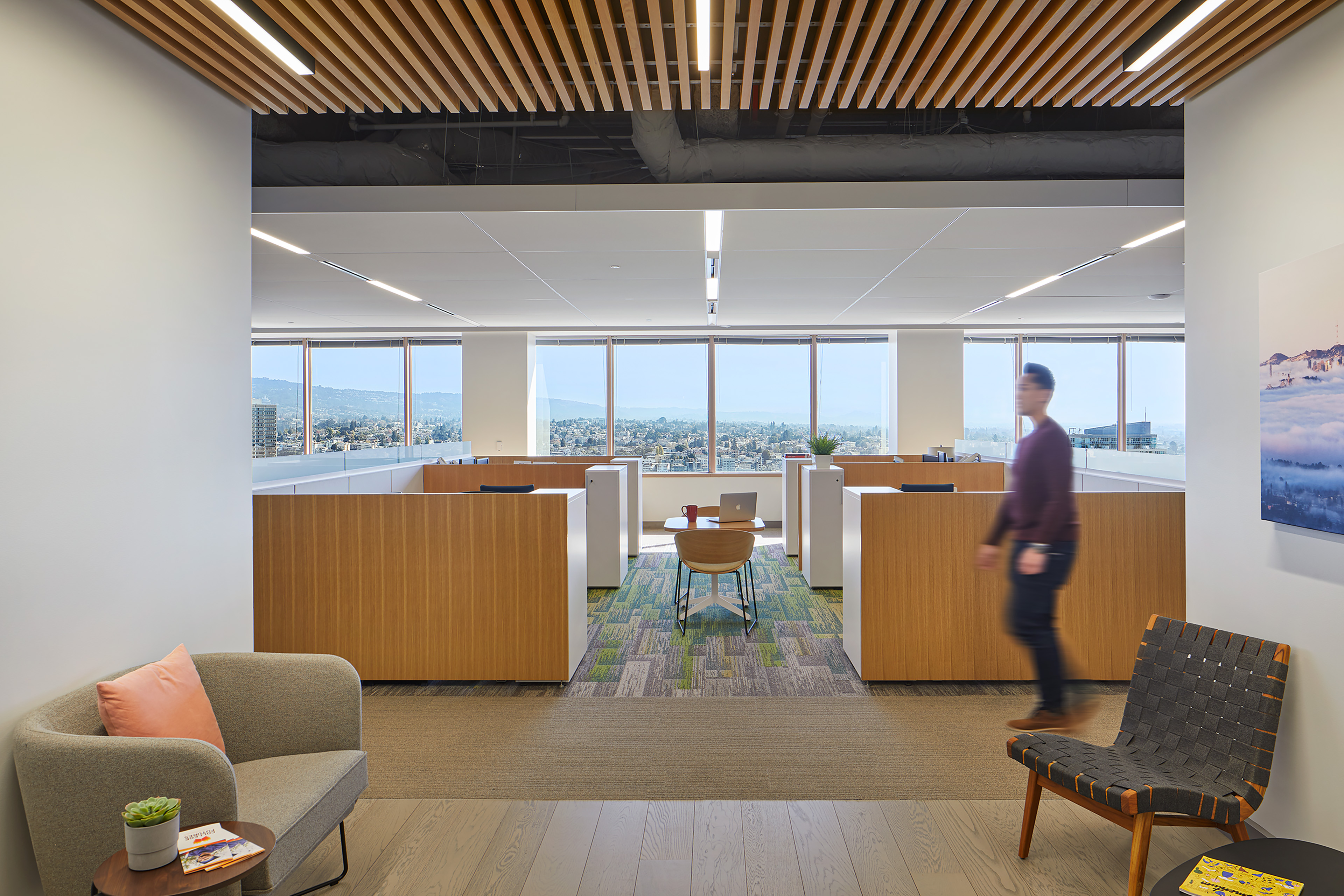 Open office entry and lounge area with wood-slat ceiling detail, soft seating, and low workstations framed by floor-to-ceiling windows with city and hillside views. A person walks through the foreground, while a central desk sits between light wood workstation panels in the bright workspace.